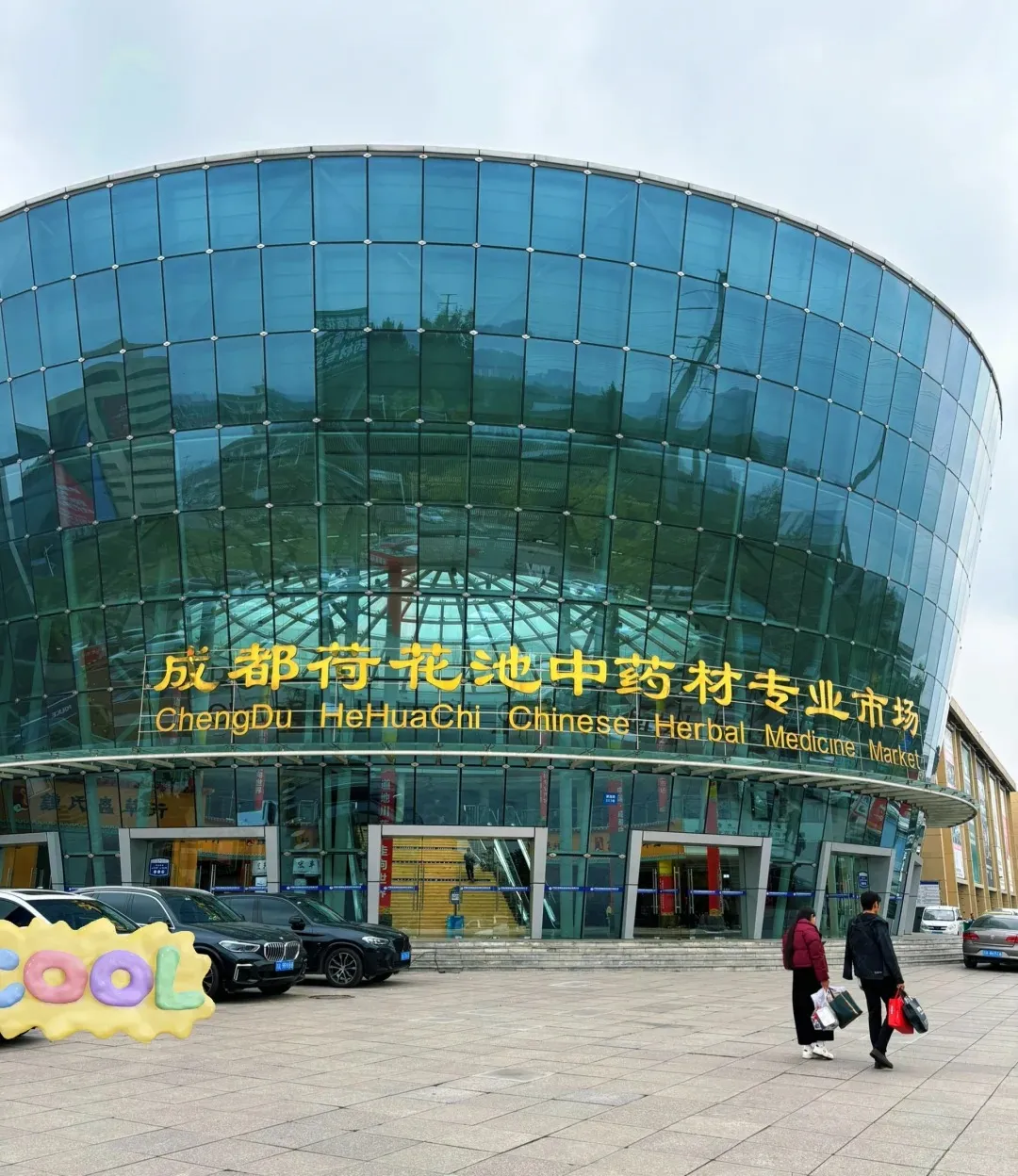 A large, modern building with a curved glass facade. Yellow Chinese characters and English text 'ChengDu HeHuaChi Chinese Herbal Medicine Market' are prominently displayed on the glass. In front, cars are parked and two people carrying bags walk across a paved plaza.