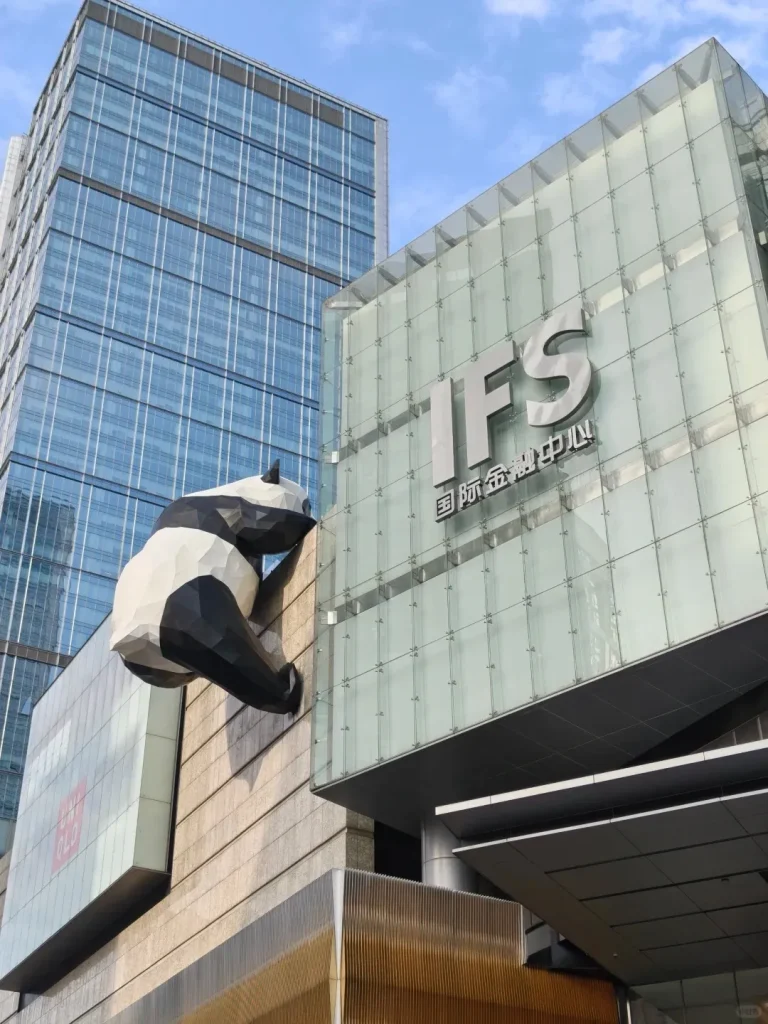 A giant, geometric black and white panda sculpture clings to the corner of a modern glass-clad building. The building prominently displays the silver 'IFS' logo and Chinese characters. In the background, another tall blue glass skyscraper rises against a partly cloudy sky, and a partial 'UNIQLO' sign is visible below the panda.
