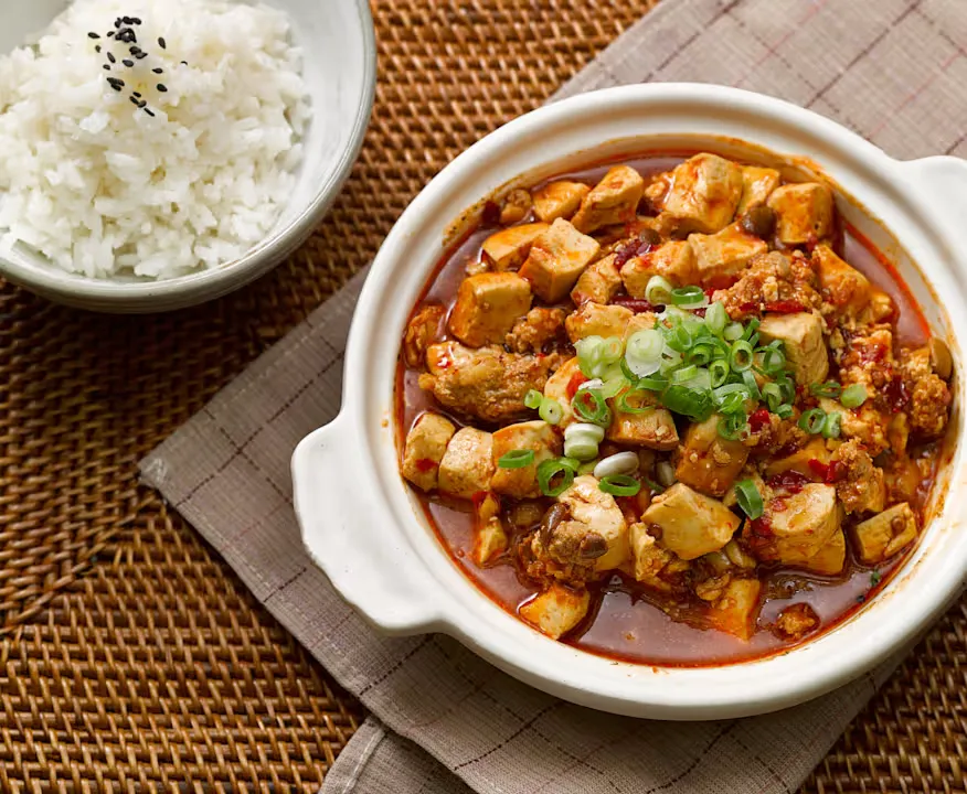 A white ceramic pot of spicy Mapo Tofu with cubed tofu, minced meat, red chili oil sauce, and green onion garnish, served next to a white bowl of white rice topped with black sesame seeds, all on a brown woven mat.