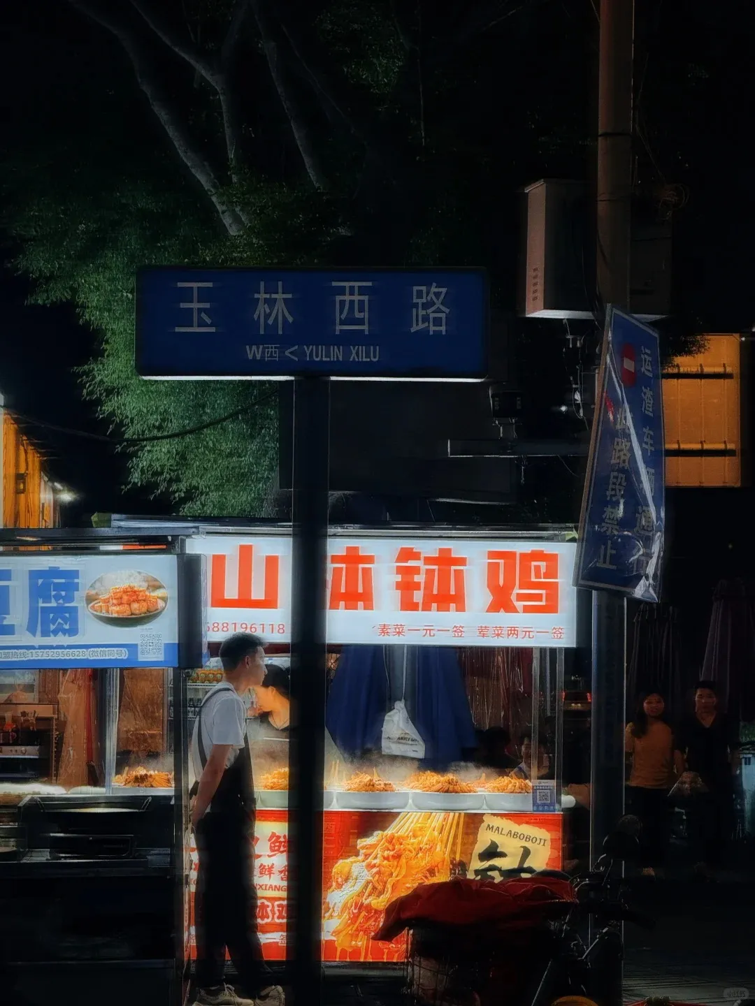 A night street scene in Chengdu, China, showing a blue street sign for 'Yulin Xilu' above two brightly lit food stalls. The stalls display various skewered foods, with a man in a white shirt and dark apron standing in front of them. In the background, dark trees and two pedestrians are visible.