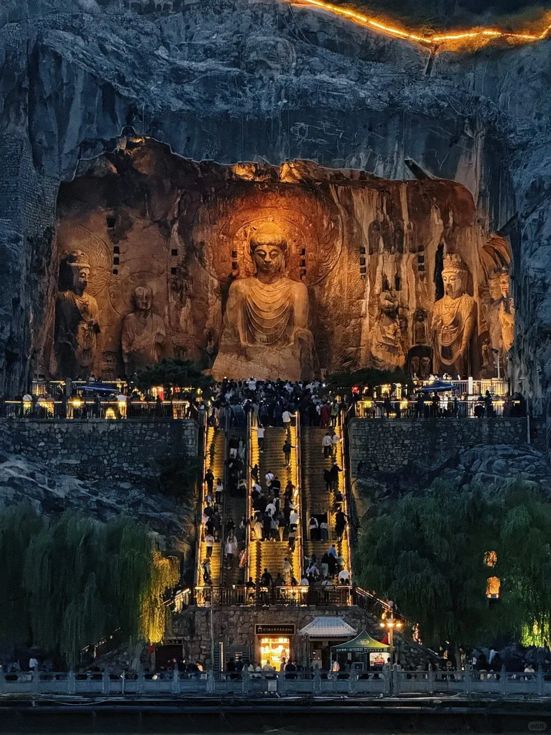 A nighttime view of the Longmen Grottoes in Luoyang, China, showing a giant seated Buddha and surrounding smaller statues carved into a rocky cliff face, all illuminated with warm golden light. Below, a wide, lit staircase filled with many visitors ascends towards the grottoes, with illuminated trees on either side.