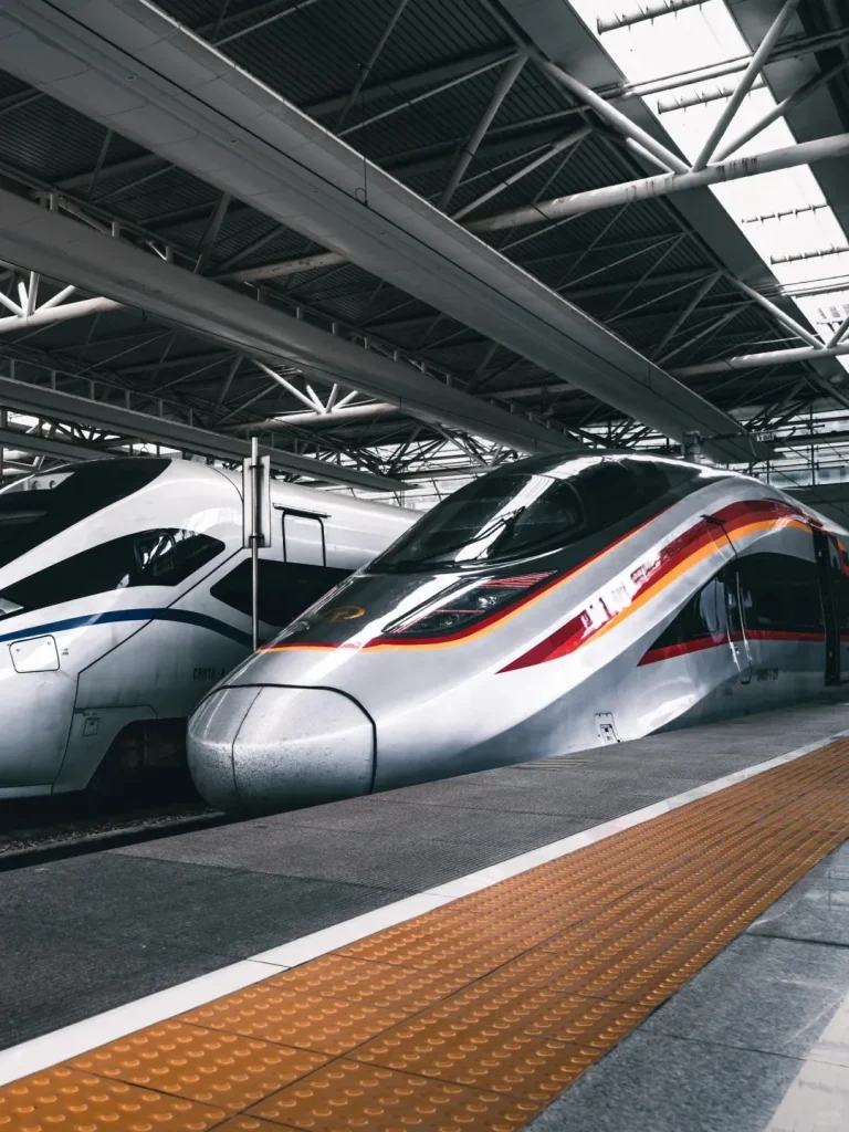 Two modern Chinese high-speed trains, including a silver Fuxing bullet train with red, orange, and gold stripes, parked side-by-side at a bright station platform. A white high-speed train with blue stripes is to the left. A yellow tactile paving strip is visible on the platform.