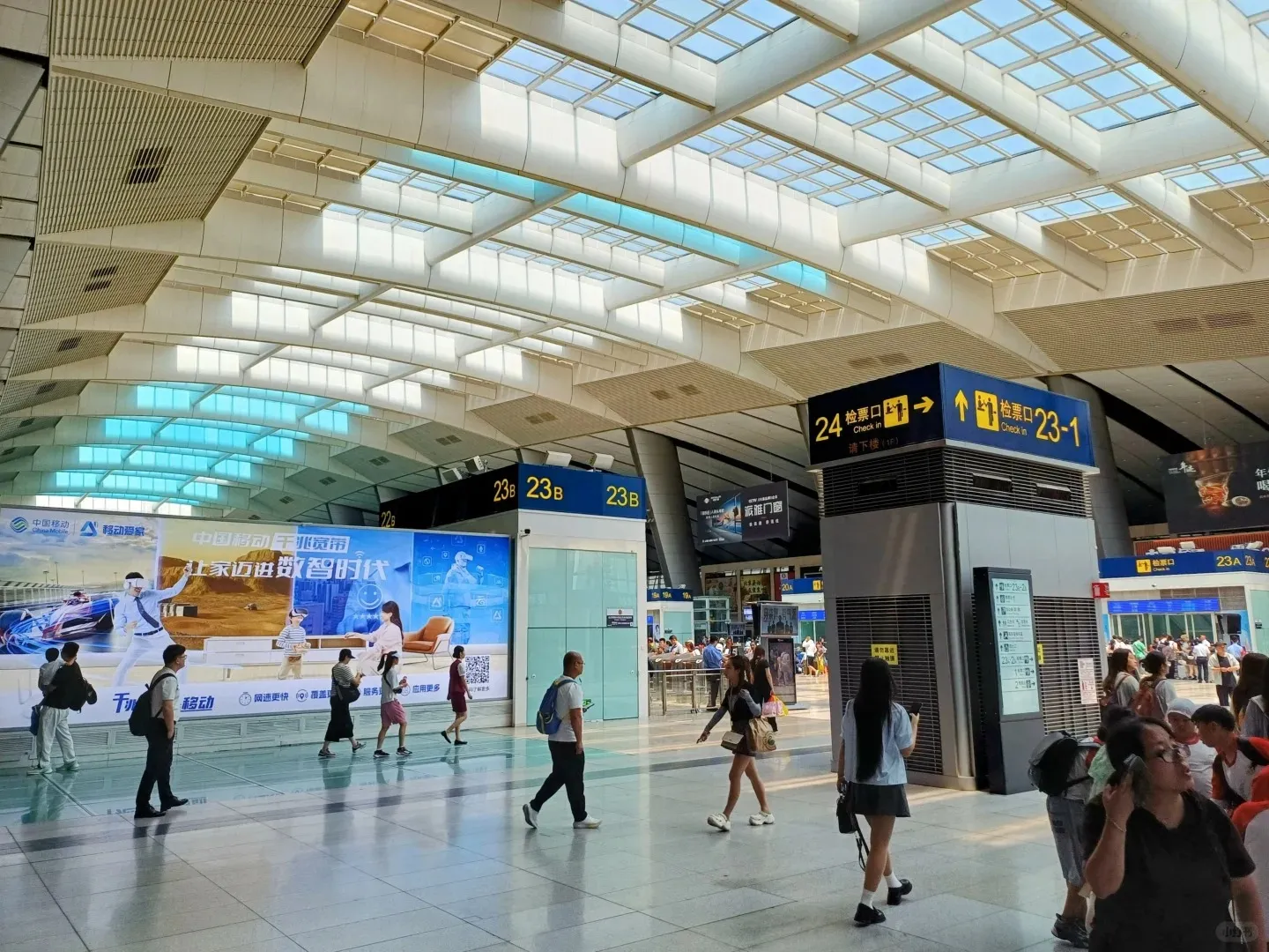 A wide interior view of a modern, brightly lit high-speed train station waiting hall. The high ceiling features skylights with light blue panels and a complex white structure. Many travelers are walking across the reflective, light-colored floor. Large digital advertisements are displayed on the far wall. A prominent blue and yellow sign directs passengers to 'Check in' gates 24 and 23-1, with an arrow pointing downstairs.