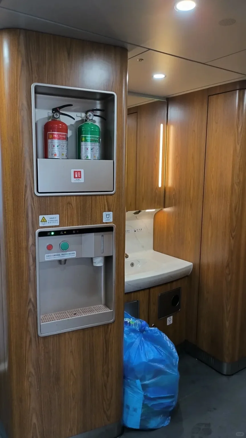 A hot water dispenser with buttons and a drip tray is integrated into a wood-paneled wall on a train. Above it, an open compartment holds a red fire extinguisher and a green fire extinguisher. To the right, a small white sink with a faucet is visible, illuminated by a vertical light strip. A blue garbage bag rests on the dark grey floor next to the sink.