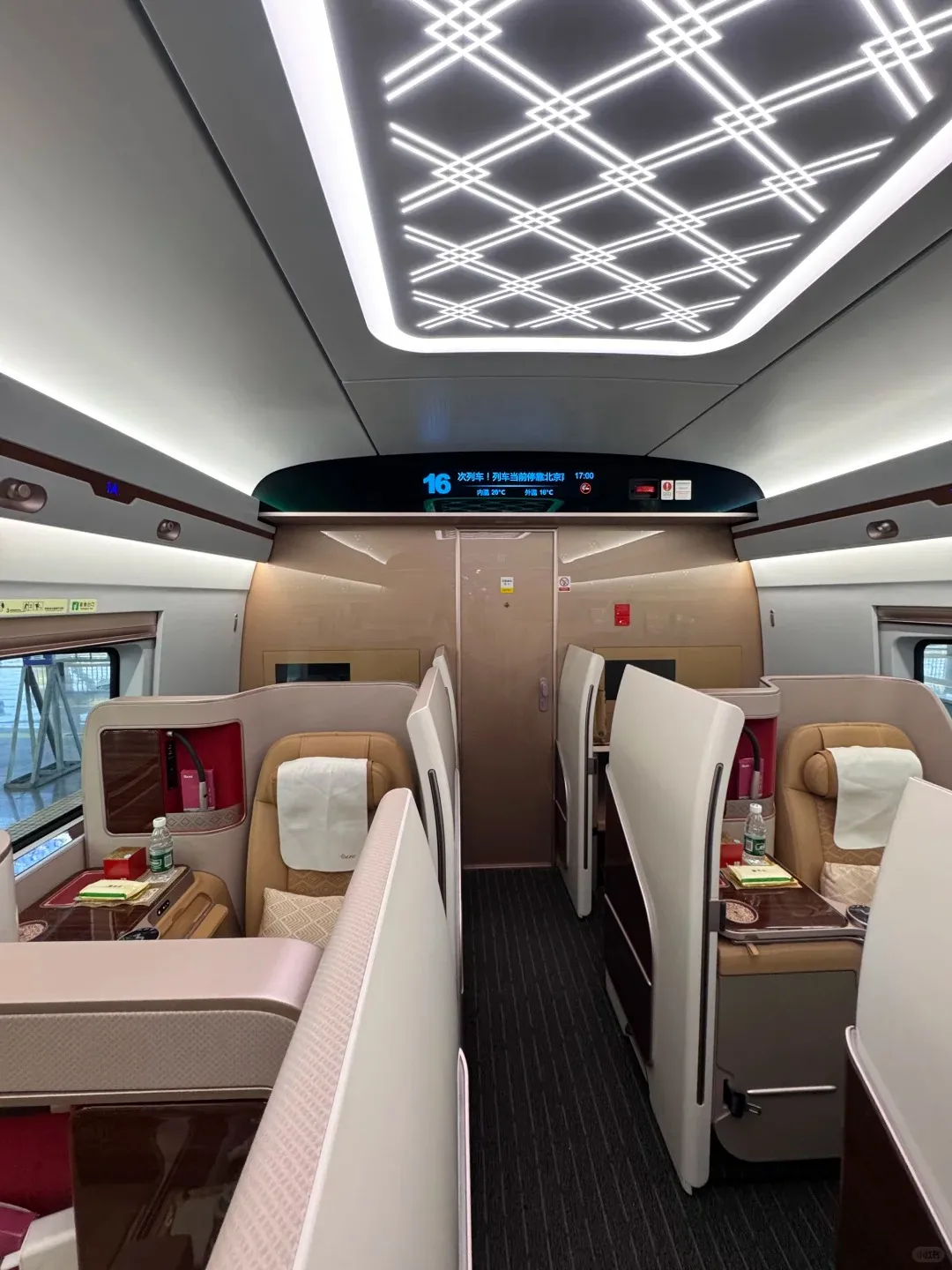 Interior view of a Chinese Fuxing high-speed train Business Class cabin, showcasing spacious individual tan leather seats with privacy dividers, small side tables with a bottle of water, and an illuminated decorative ceiling. A digital display board at the front indicates the train is stopped at Beihai, with an internal temperature of 20°C and external temperature of 16°C.