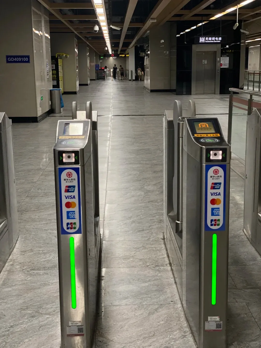 Two modern silver subway turnstiles with glowing green indicator lights stand prominently in the foreground. The turnstiles display accepted payment methods including e-CNY, UnionPay, Visa, American Express, and JCB. In the background, a wide, brightly lit metro station hallway stretches into the distance with a few commuters and an accessible elevator sign.