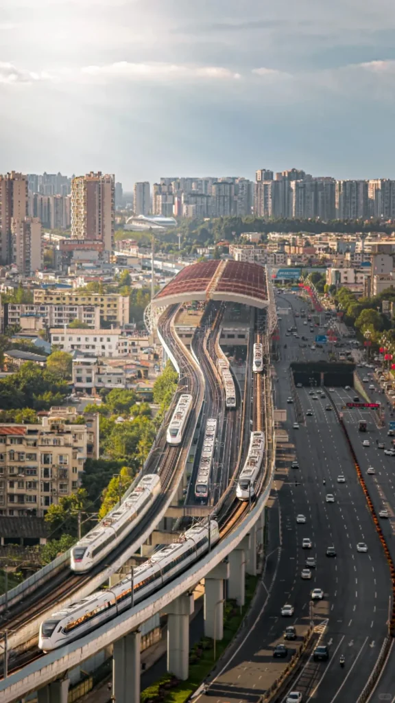 An aerial perspective of a modern Chinese city showcasing multiple elevated train tracks with several white trains, converging at a large station with an arched reddish-brown roof. A multi-lane highway runs parallel to the rail lines, bustling with cars. In the background, numerous high-rise buildings fill the urban skyline.