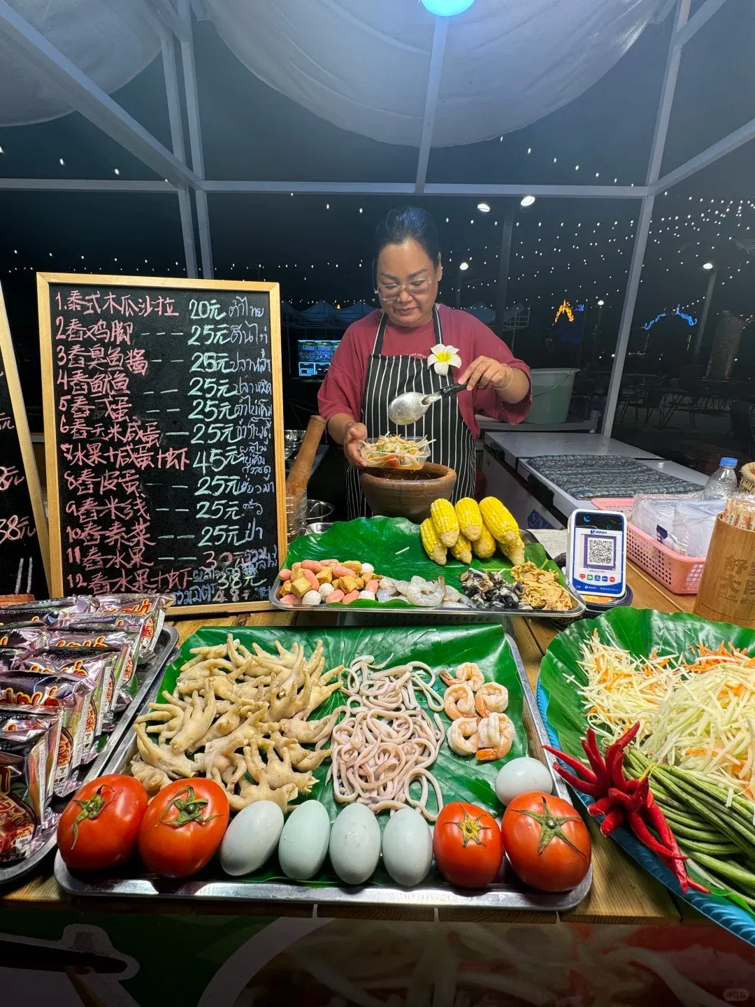 A street food vendor in a striped apron scoops food from a wooden mortar into a bowl at a bustling night market stall. Trays of fresh ingredients including chicken feet, squid, shrimp, blue duck eggs, tomatoes, corn cobs, and various chopped vegetables like papaya, long beans, and chilies are displayed. A blackboard menu with Chinese and Thai text and a mobile payment QR code are visible on the counter.