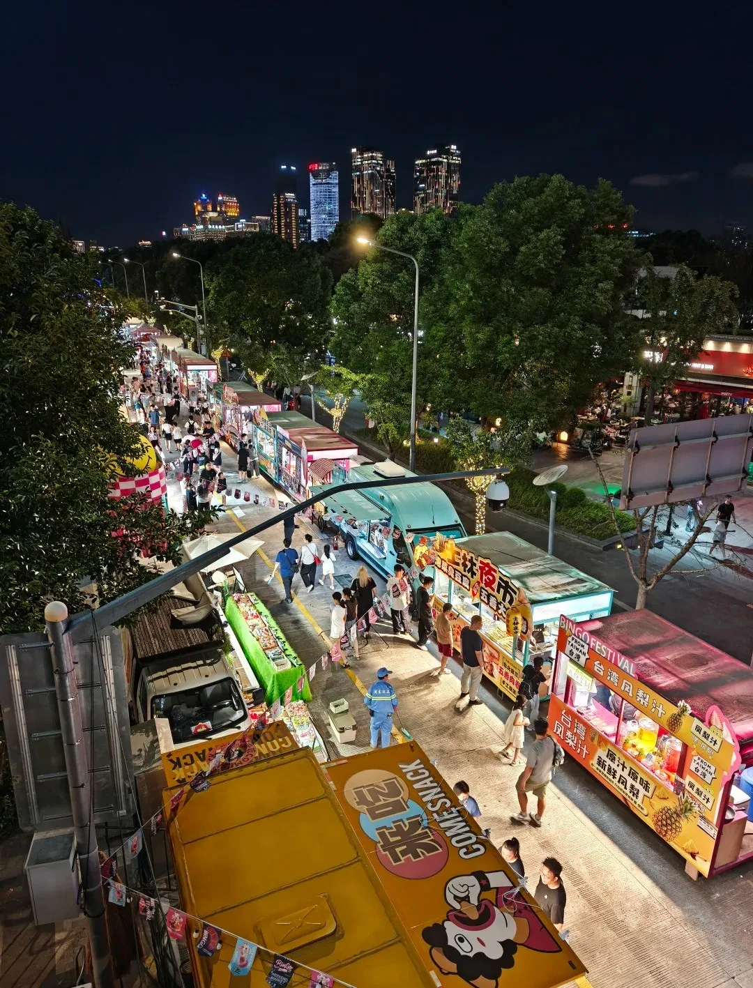 High-angle view of a bustling night market street in a city, lined with numerous brightly lit food trucks and stalls. Many people are walking, browsing, and buying food. Tall, illuminated buildings rise in the background against a dark night sky, with trees along the street. A large yellow food truck in the foreground reads 'COME SNACK' and a pink one behind it advertises 'BINGO FESTIVAL' and 'FROM TAIWAN' with fruit drinks.