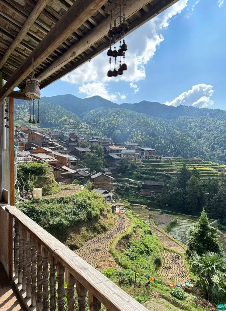 View from a traditional wooden guesthouse balcony overlooking a rural village built into green mountains, with terraced rice paddies and cultivated fields below. Decorative wooden bells hang from the visible wooden ceiling beams, under a blue sky with white clouds.