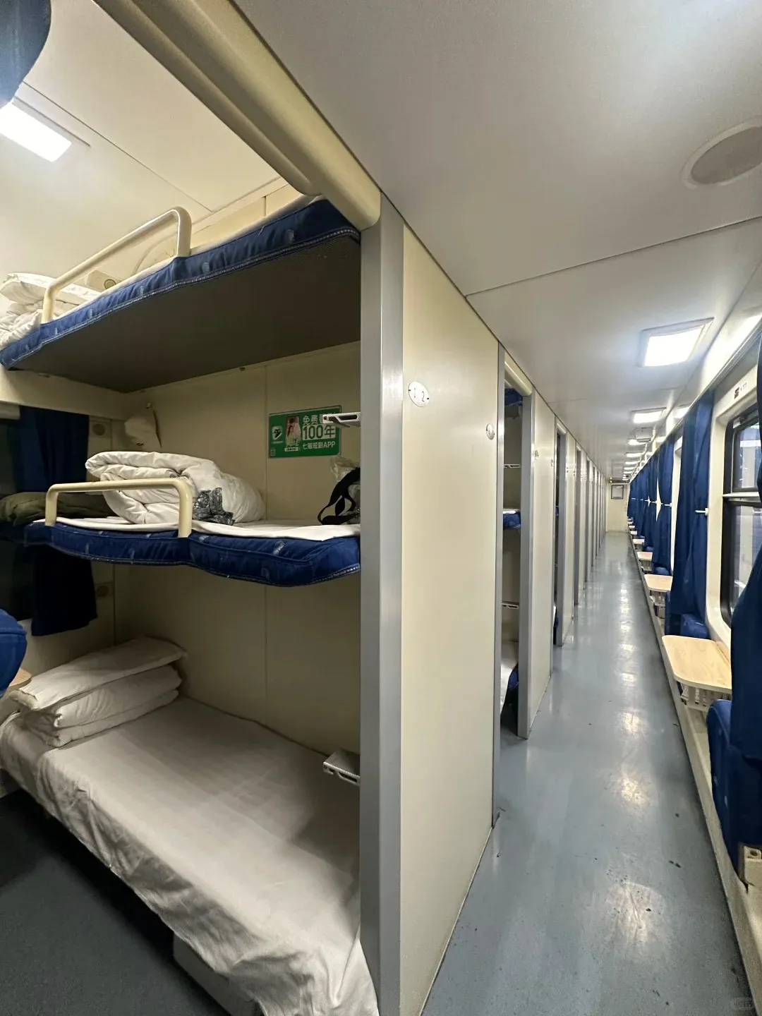 Interior view of a Chinese hard sleeper train car. On the left, a bay with three bunk beds is visible: a made-up lower berth, a middle berth with a blue mattress and bedding, and an upper berth with a blue mattress. A narrow corridor stretches into the distance, flanked by more sleeping berths on the left and windows with blue curtains and small wooden tables on the right.