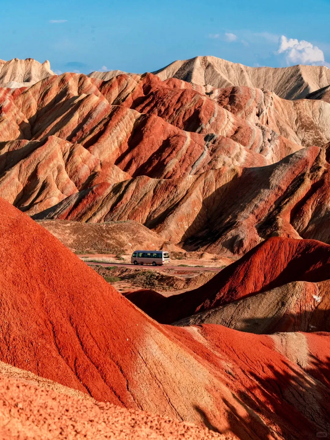 A vast landscape of vibrant, layered Danxia mountains, featuring alternating stripes of deep red, orange, yellow, and beige rock formations. A white tour van travels on a winding road through the hilly terrain under a clear blue sky.