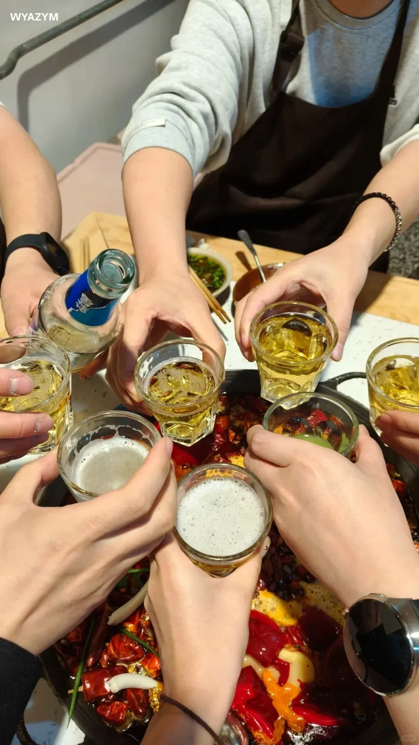 A group of people's hands hold small glasses filled with beer and a beer bottle, raised in a toast over a bubbling hot pot rich with red chili peppers and other ingredients.