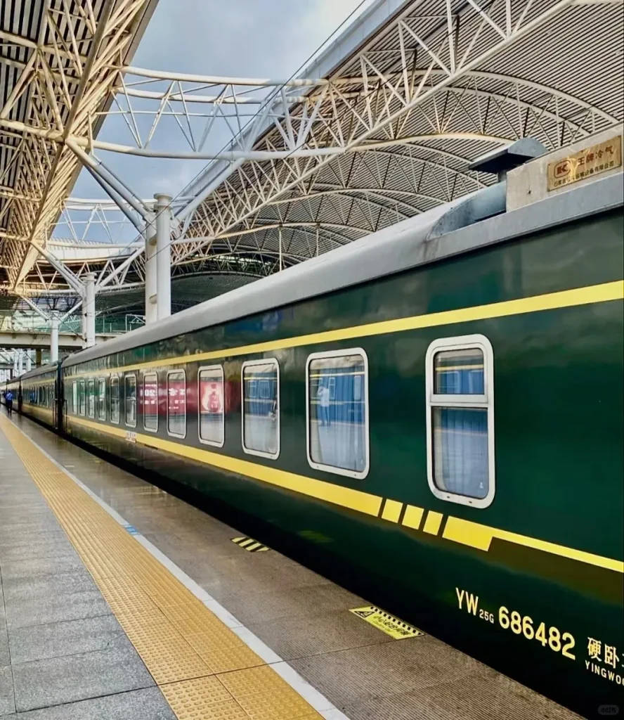 A dark green Chinese train car with a prominent yellow stripe is parked at a station platform. The train windows reflect the overhead white metal truss roof structure of the station. Yellow textured safety strips run along the platform edge. Text on the train car reads 'YW 25G 686482 硬卧'.