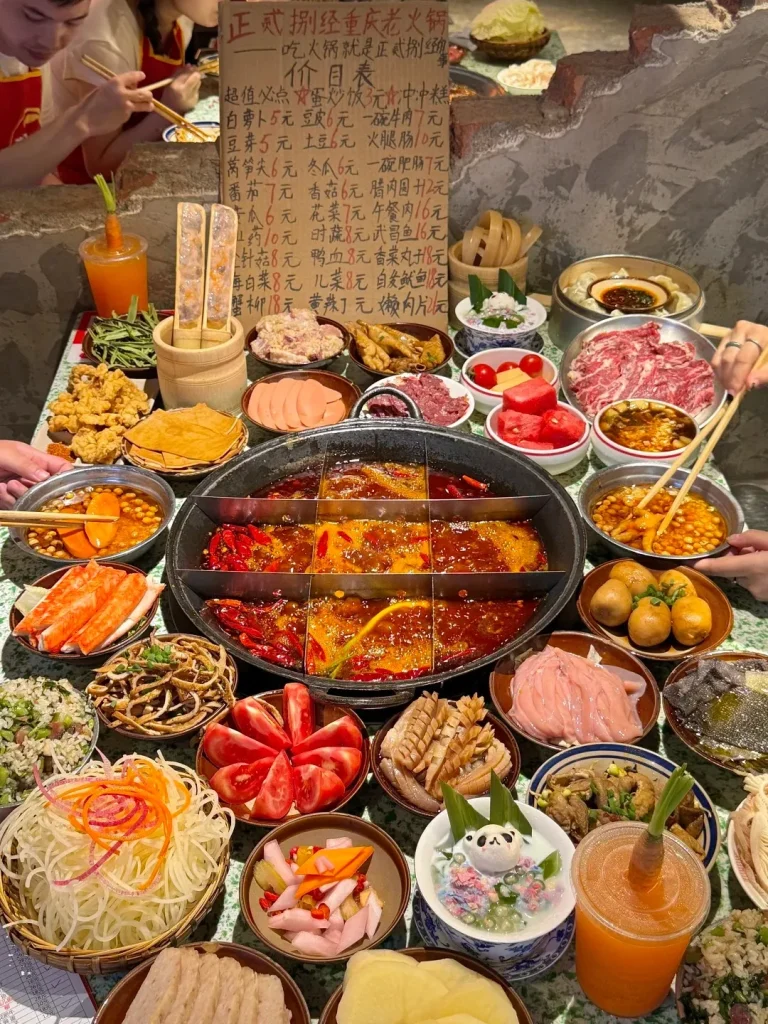 A vibrant overhead view of a large, multi-compartment Chinese hot pot on a table, filled with spicy red broth and surrounded by numerous small bowls and plates of raw ingredients, cooked side dishes, dipping sauces, and drinks. Multiple hands are visible using chopsticks to pick up food from the communal spread.