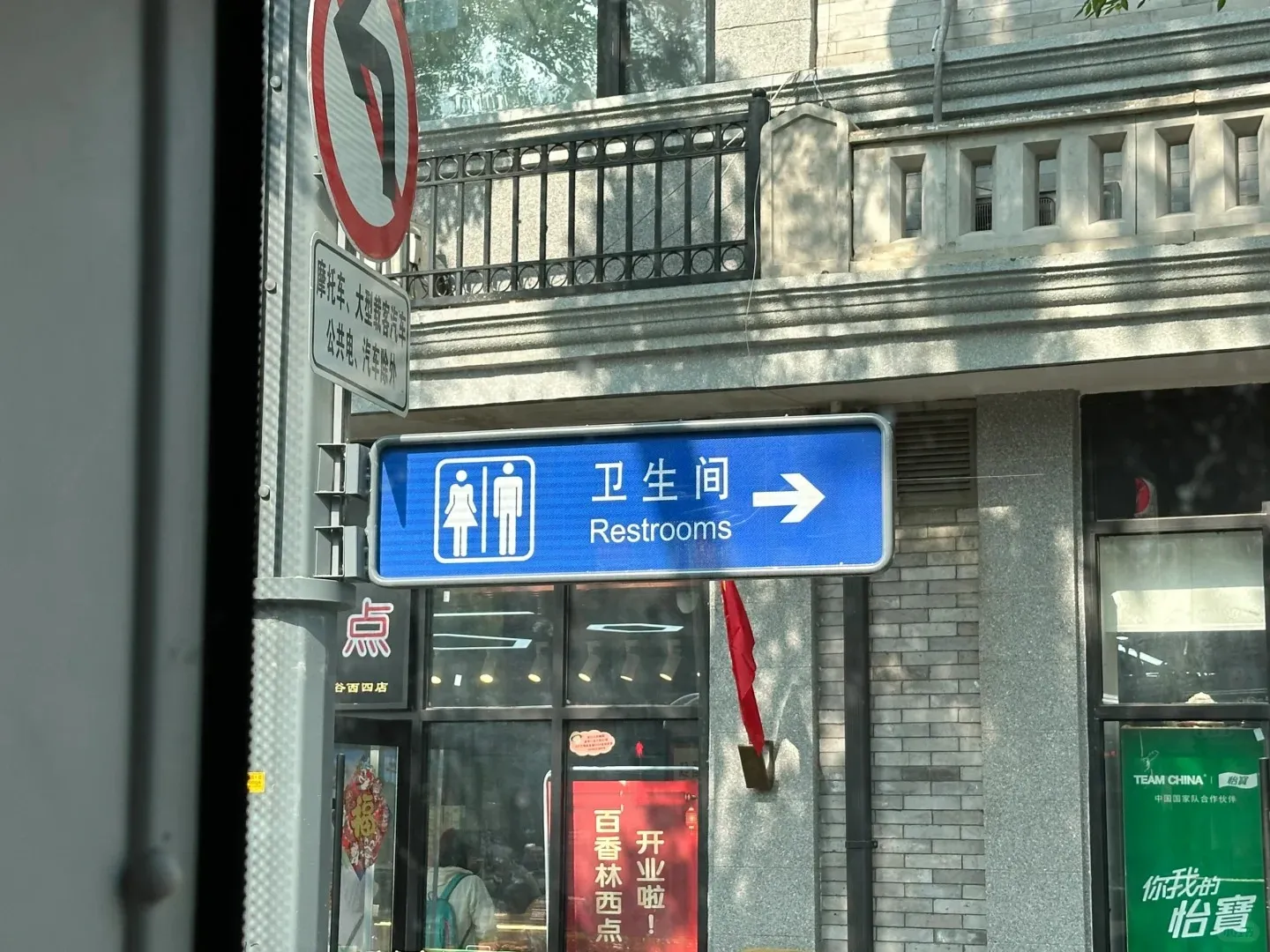 A blue rectangular public restroom sign featuring white icons of a woman and a man, Chinese characters '卫生间', and the English word 'Restrooms' with a white arrow pointing right. The sign is attached to a grey pole, with a partially visible red circular 'no left turn' traffic sign above it, and a stone building facade in the background.