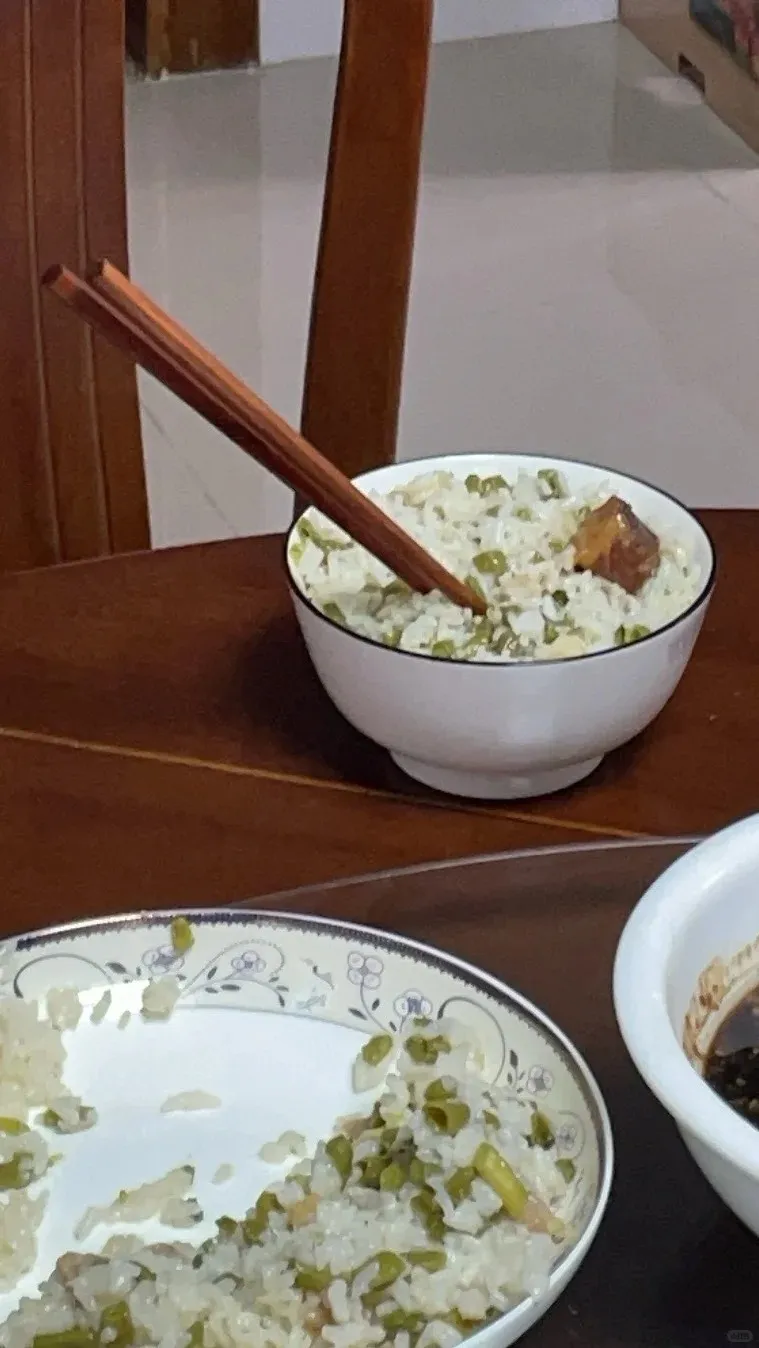 A pair of brown wooden chopsticks are stuck vertically into a white bowl of rice mixed with green peas and a small piece of meat. The bowl sits on a dark wooden table, with a partially eaten plate of similar rice visible in the foreground.