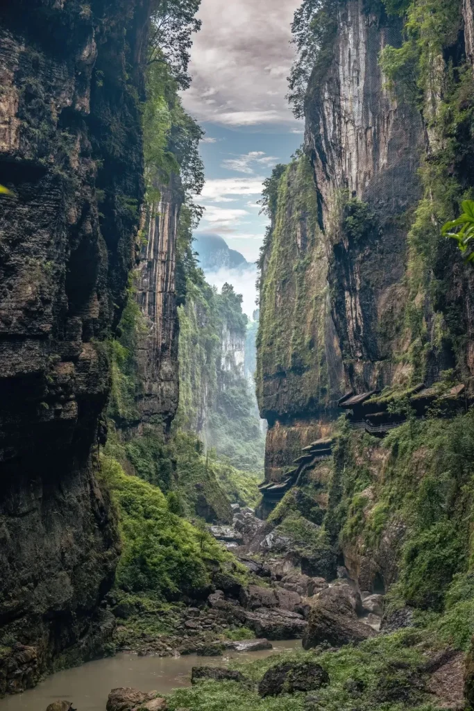 A deep, narrow canyon with towering, green-vegetated limestone cliffs. A winding wooden walkway with traditional-style roofs is built into the right cliff face. A rocky riverbed with a muddy stream runs through the bottom of the gorge. Distant mountains are shrouded in mist under a cloudy sky.