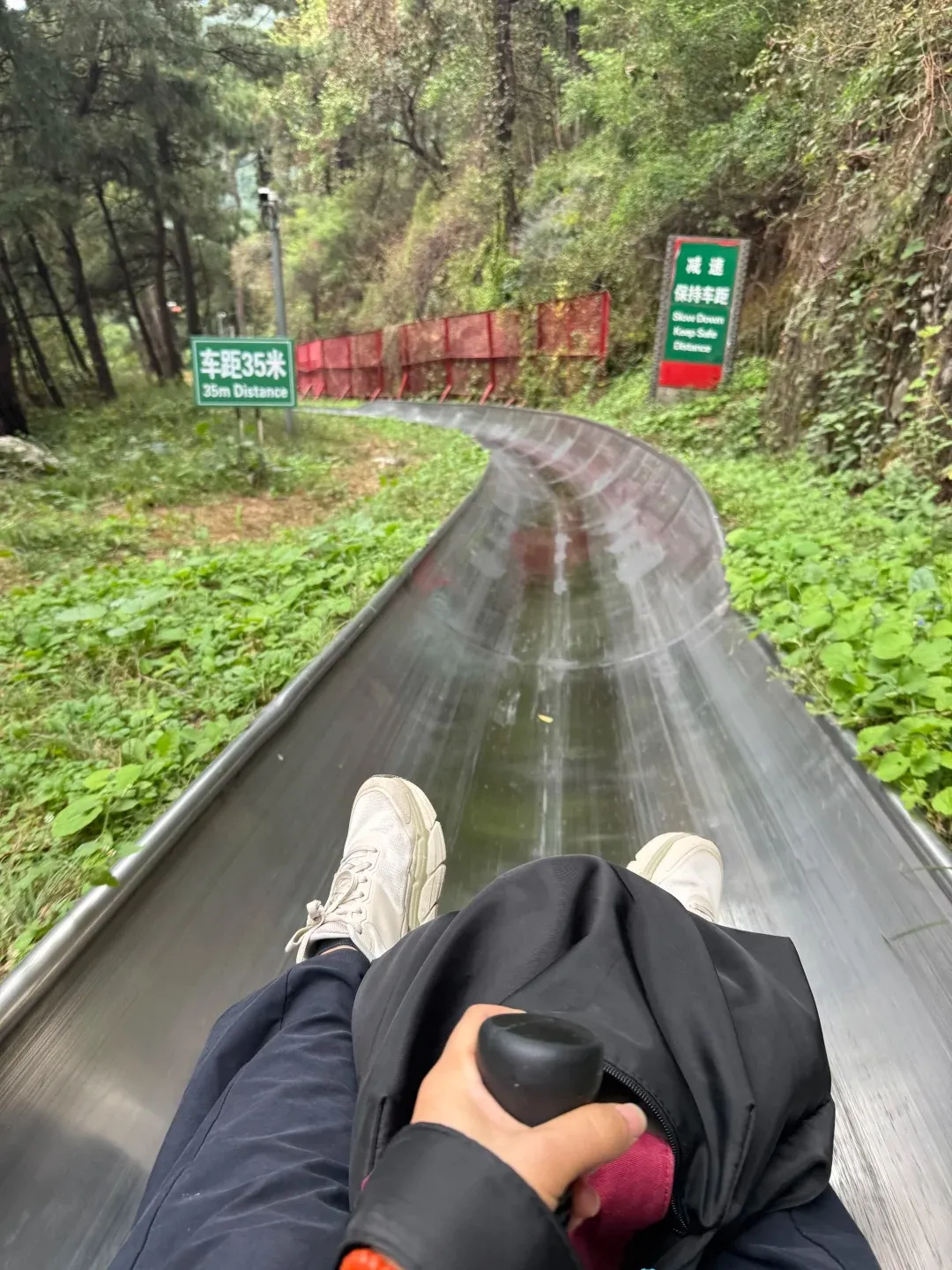 First-person perspective of a person riding a metal toboggan slide, a popular way to descend from the Great Wall of China. The rider's legs in dark pants and white sneakers are visible, along with a hand holding the black brake handle. The wet, shiny track curves through a lush green forest, with safety signs in Chinese and English ('35m Distance', 'Slow Down, Keep Safe Distance') visible on the side.