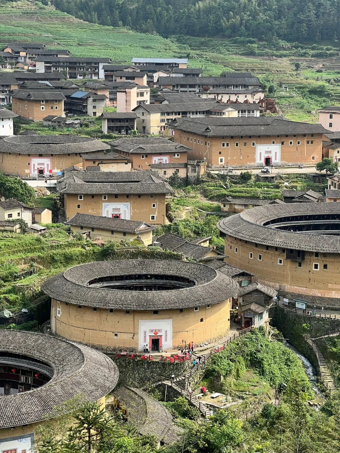 High-angle aerial view of a Fujian Tulou cluster, showcasing several large, round and square earthen buildings with dark tiled roofs. The distinctive light brown structures are nestled amidst lush green terraced hillsides and smaller traditional houses, with winding pathways connecting them.
