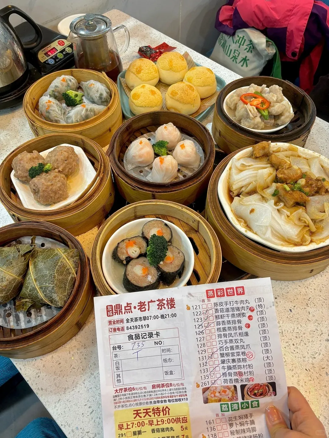 A table in a teahouse laden with a variety of Cantonese dim sum dishes. Bamboo steamers contain shrimp dumplings, other steamed dumplings, steamed meatballs, and chicken feet. Plates hold golden pineapple buns, rice noodle rolls with pork ribs, lotus leaf-wrapped sticky rice, and seaweed-wrapped rolls. A teapot and a restaurant order card are also visible.