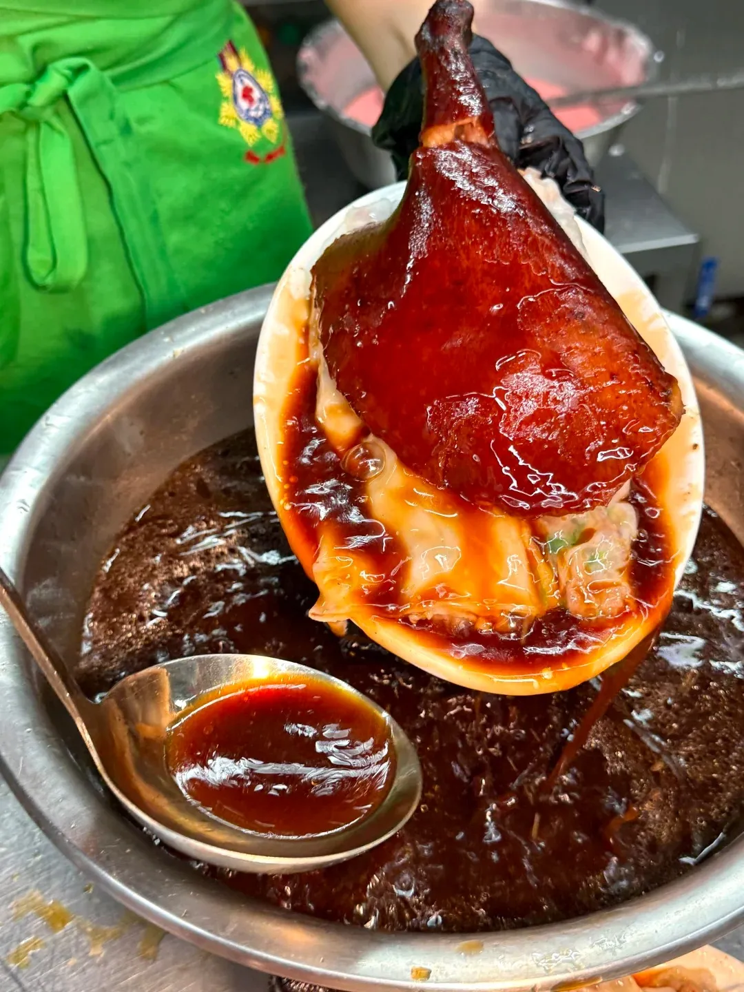 A roasted goose leg with glistening reddish-brown skin is held by a hand in a black glove, dripping dark brown sauce into a large metal bowl of more sauce. White dumplings are visible under the leg on a plate. A metal ladle filled with sauce is in the bowl.