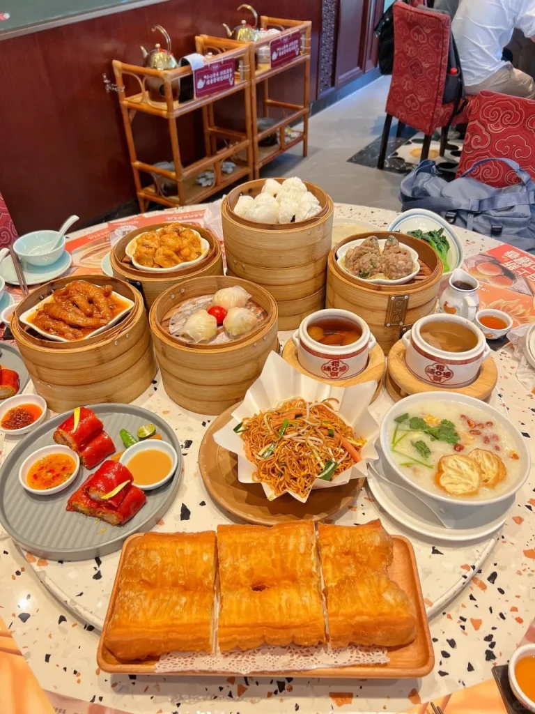 A large round table in a traditional Cantonese restaurant displaying a wide array of dim sum dishes, including multiple bamboo steamers with various dumplings and buns, a plate of red glutinous rice rolls, fried noodles, congee with fried dough sticks (youtiao), and small bowls of braised dishes.