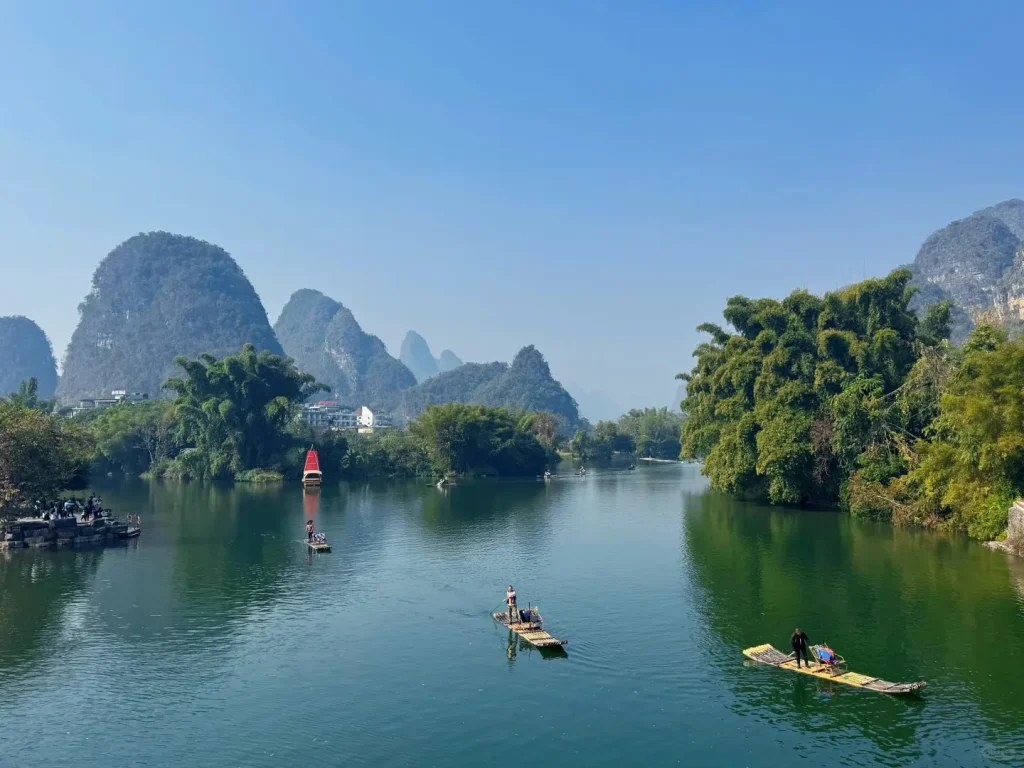 Several bamboo rafts with people float on a wide, green river. The river is surrounded by dense green trees and towering, rounded karst limestone mountains under a clear blue sky. On the left bank, a stone pier holds onlookers, and a decorative red boat is anchored further downriver near some buildings.