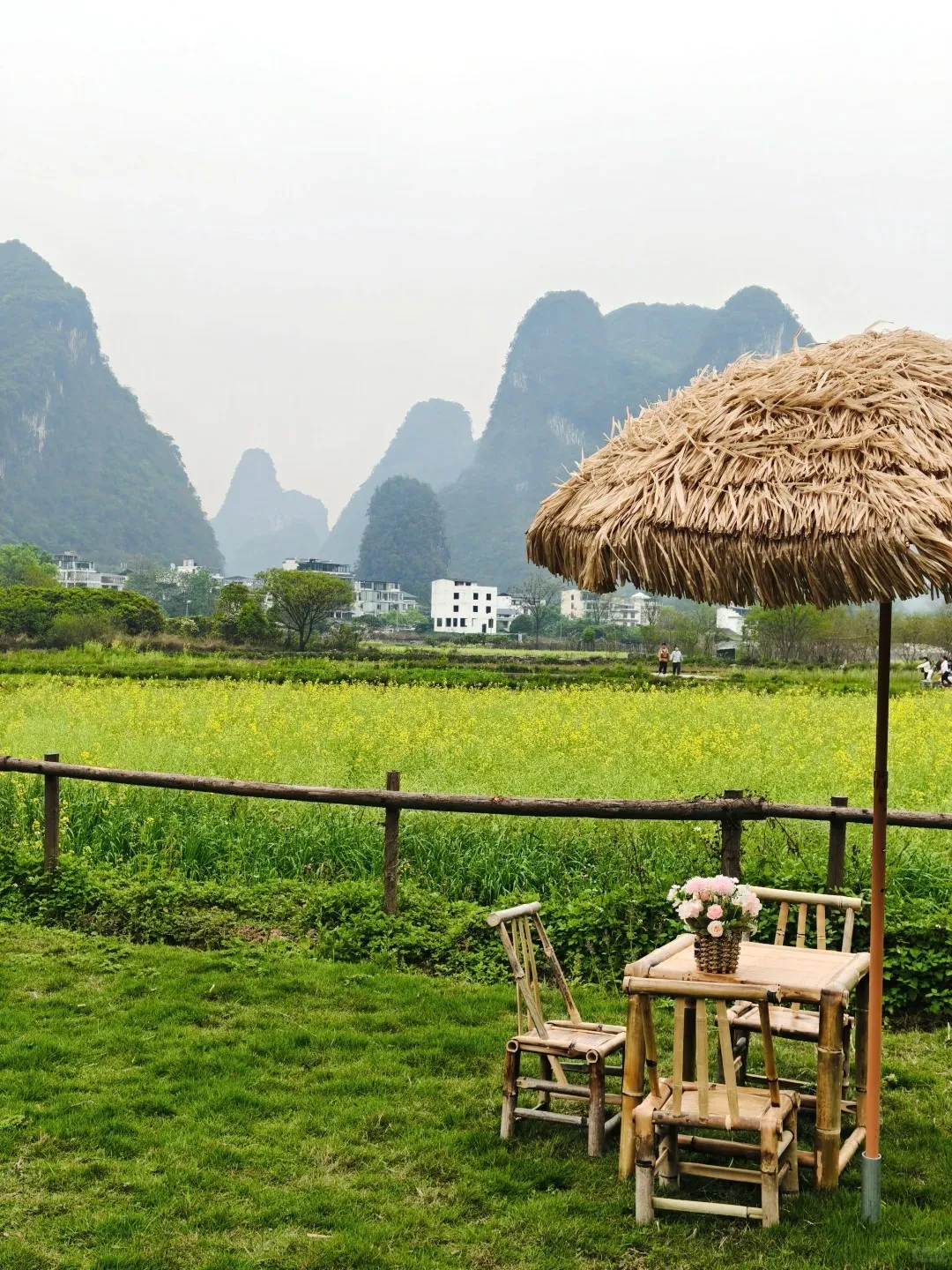 A scenic view with a green grassy lawn in the foreground, featuring a rustic bamboo table, two bamboo chairs, and a straw umbrella. A small woven basket with pink flowers sits on the table. Behind a low wooden fence, a vibrant yellow field of rapeseed flowers stretches towards the midground. In the distance, majestic karst mountains rise under a hazy sky, with white buildings nestled at their base.