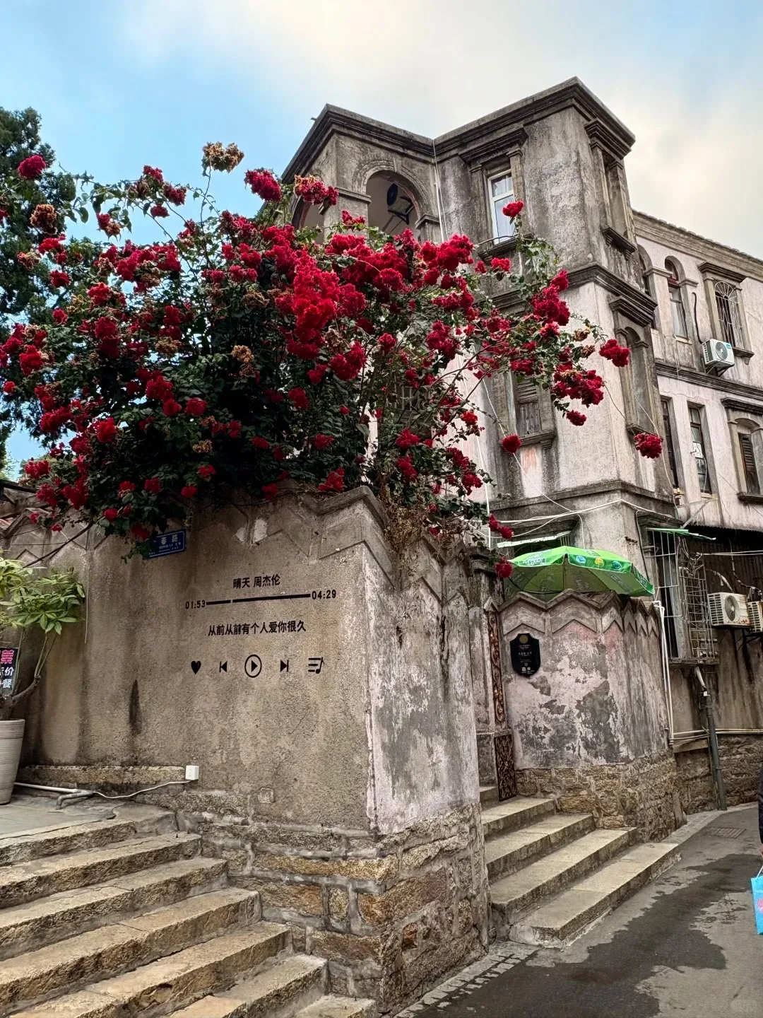 A weathered, grey colonial-style building on Gulangyu Island featuring a prominent tower, arched balcony, and multiple windows. A large bush of bright red flowers covers the lower front. Stone steps ascend from the street on both sides. On the adjacent concrete wall, black painted text includes Chinese characters, timestamps '01:53 - 04:29', and music player symbols like a heart and play button.