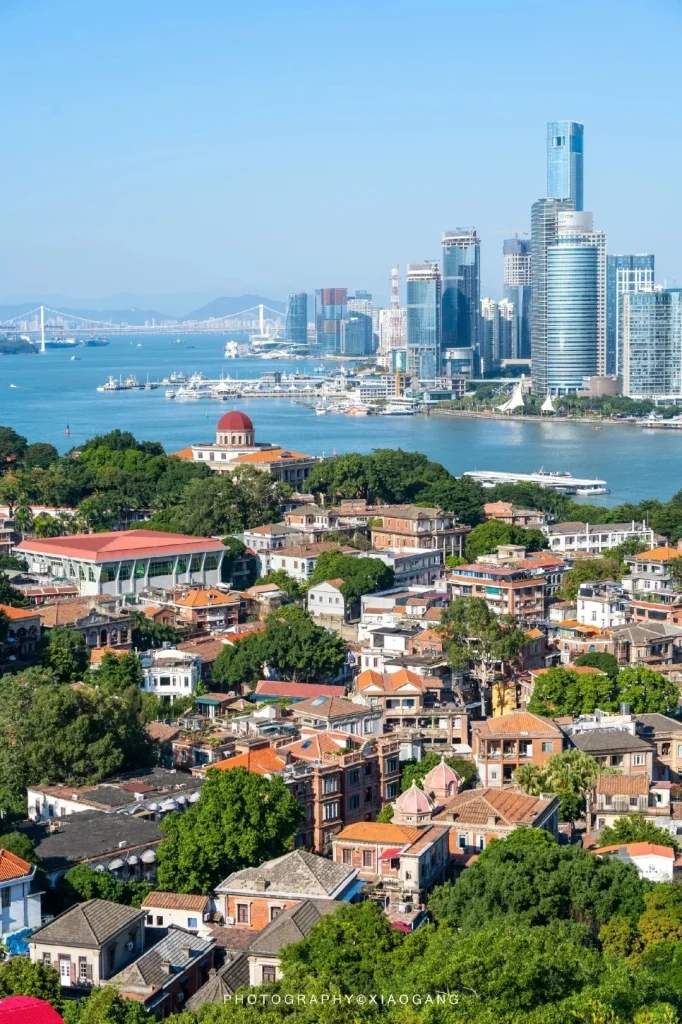 Elevated view of Gulangyu Island showing historic European-style buildings with red roofs, surrounded by dense green trees. In the middle ground, boats dot the sparkling blue water, leading to the modern Xiamen city skyline in the background, featuring numerous tall glass skyscrapers and a long suspension bridge.