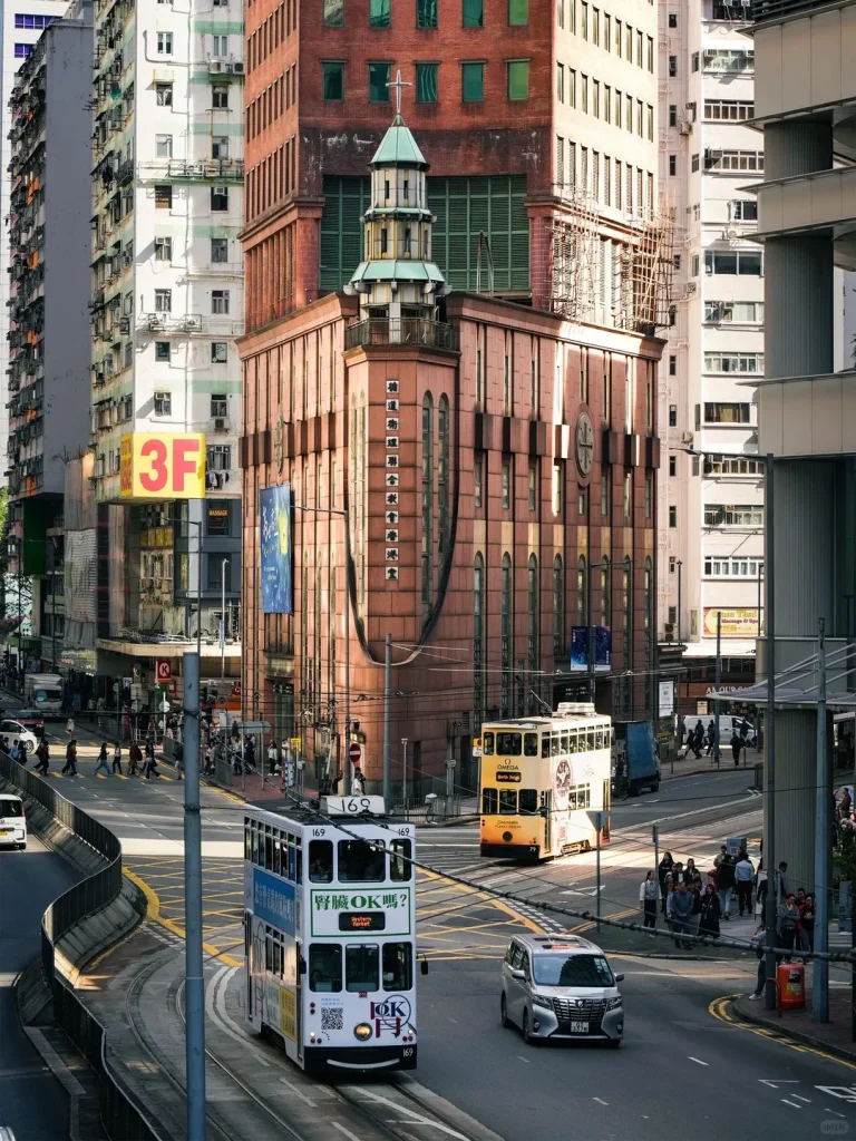 A bustling street scene in Hong Kong features a historic peach-colored building with a green spire and cross, flanked by modern high-rises. Two iconic double-decker trams, one white and blue (number 169) and one yellow (number 79), are visible on the tracks. Pedestrians, cars, and other vehicles fill the busy intersection.
