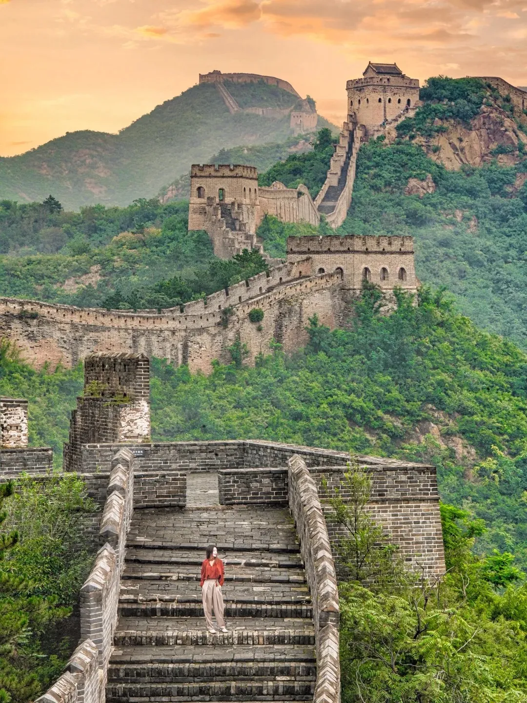The Jinshanling section of the Great Wall of China winds along lush green mountain ridges, featuring multiple ancient brick watchtowers under a warm orange sunset sky. A person in a red shirt and light pants stands on the wall's steps in the foreground.