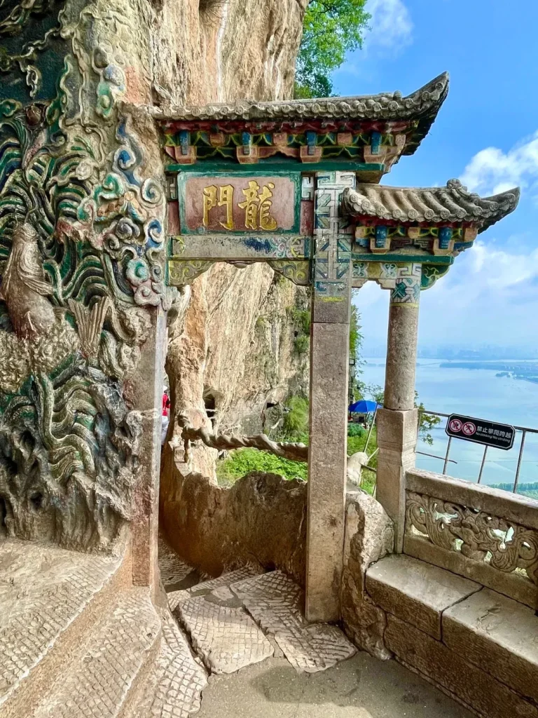 An ornate traditional Chinese archway, known as the Dragon Gate, is carved into a rugged cliff face. The archway features a colorful tiered roof and a green plaque with the gold Chinese characters '龍門' (Dragon Gate). Intricate stone carvings of a dragon and swirling patterns adorn the cliff to the left. Through the archway, a vast blue Dianchi Lake and a distant city skyline are visible under a blue, partly cloudy sky. A carved stone railing and bench are to the right, along with a 'No climbing/No striding' sign.