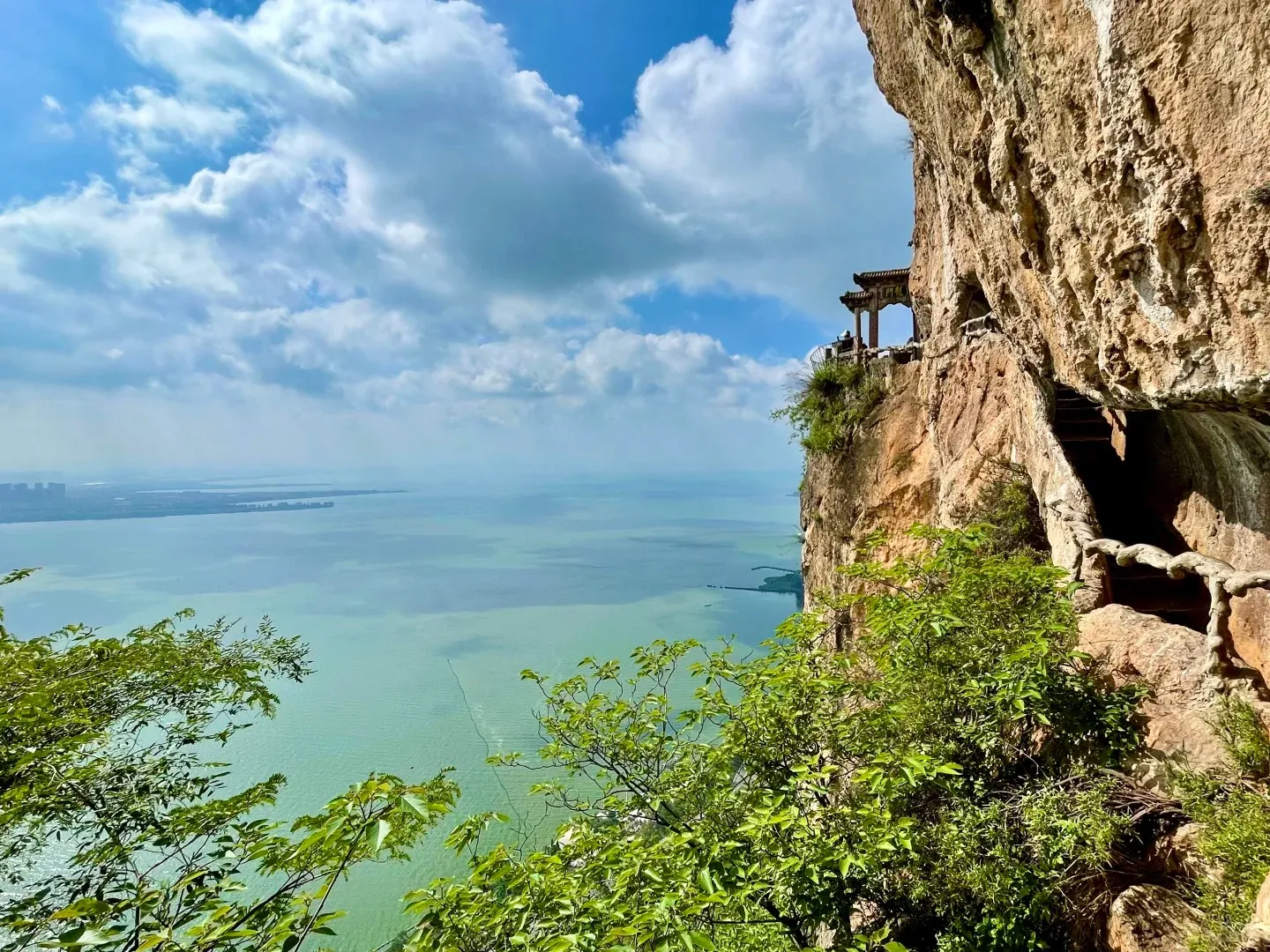 A panoramic view from a rugged cliff showing a traditional Chinese pavilion built on the edge, with carved stone steps leading into grottoes. Below, the vast, turquoise Dianchi Lake extends to the horizon, with distant city buildings visible on the far shore under a blue sky with white clouds. Green foliage frames the lower foreground.
