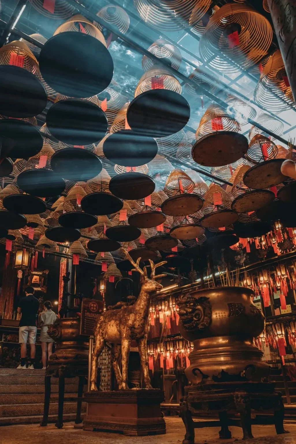 Interior of Man Mo Temple in Hong Kong, featuring hundreds of large spiral incense coils with red tags hanging from the ceiling. Sunlight beams through the smoke, illuminating a golden deer statue and a large ornate bronze incense censer in the foreground, with two people in the background.