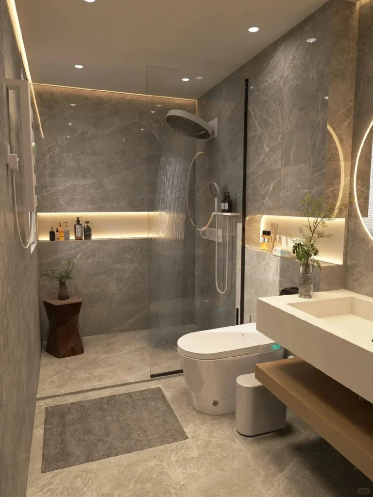 A modern bathroom featuring grey marble-patterned tiles on the walls and floor. On the left, a walk-in shower with a glass partition and a rainfall showerhead actively spraying water. In the center, a white Western-style smart toilet is positioned next to a small white waste bin. To the right, a floating white vanity with a rectangular sink and a wooden shelf underneath. The bathroom is illuminated by recessed ceiling lights, LED strip lighting in wall niches, and a circular backlit mirror. Various toiletries are arranged on the shelves.