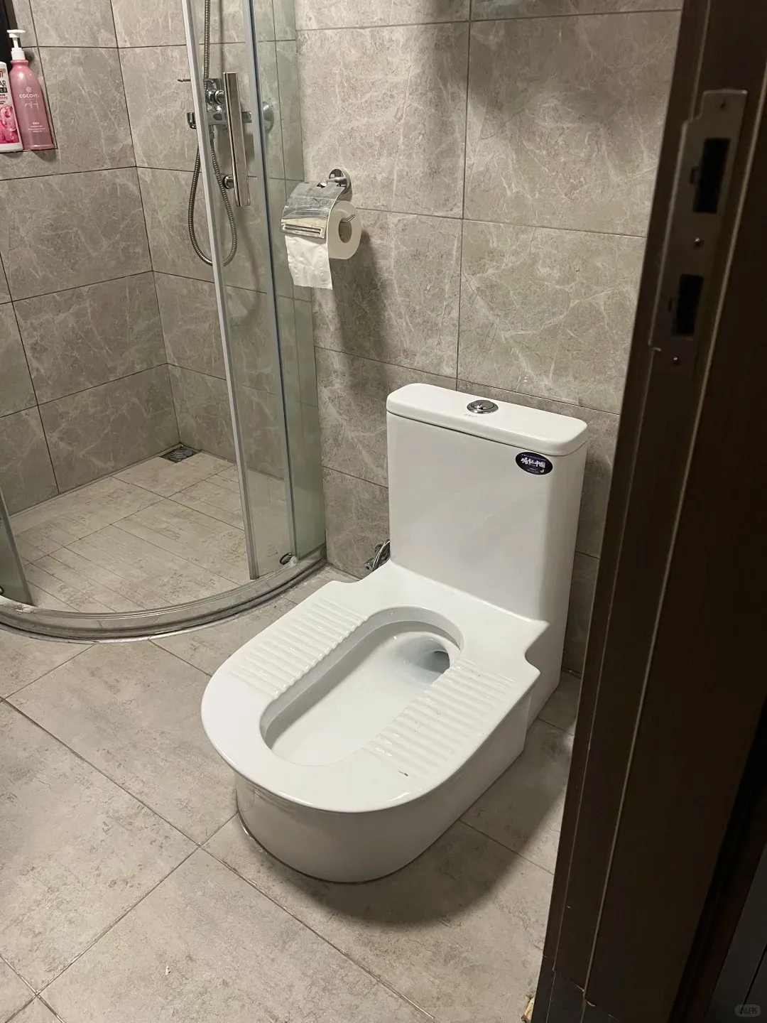 A modern white elevated squat toilet with a visible tank, featuring textured anti-slip grooves on its footrests. The toilet is positioned in a clean bathroom with light grey marble-patterned wall and floor tiles. To the left, a glass shower enclosure with a shower head and bottles of toiletries is visible. A chrome toilet paper holder with a roll of paper is mounted on the tiled wall.