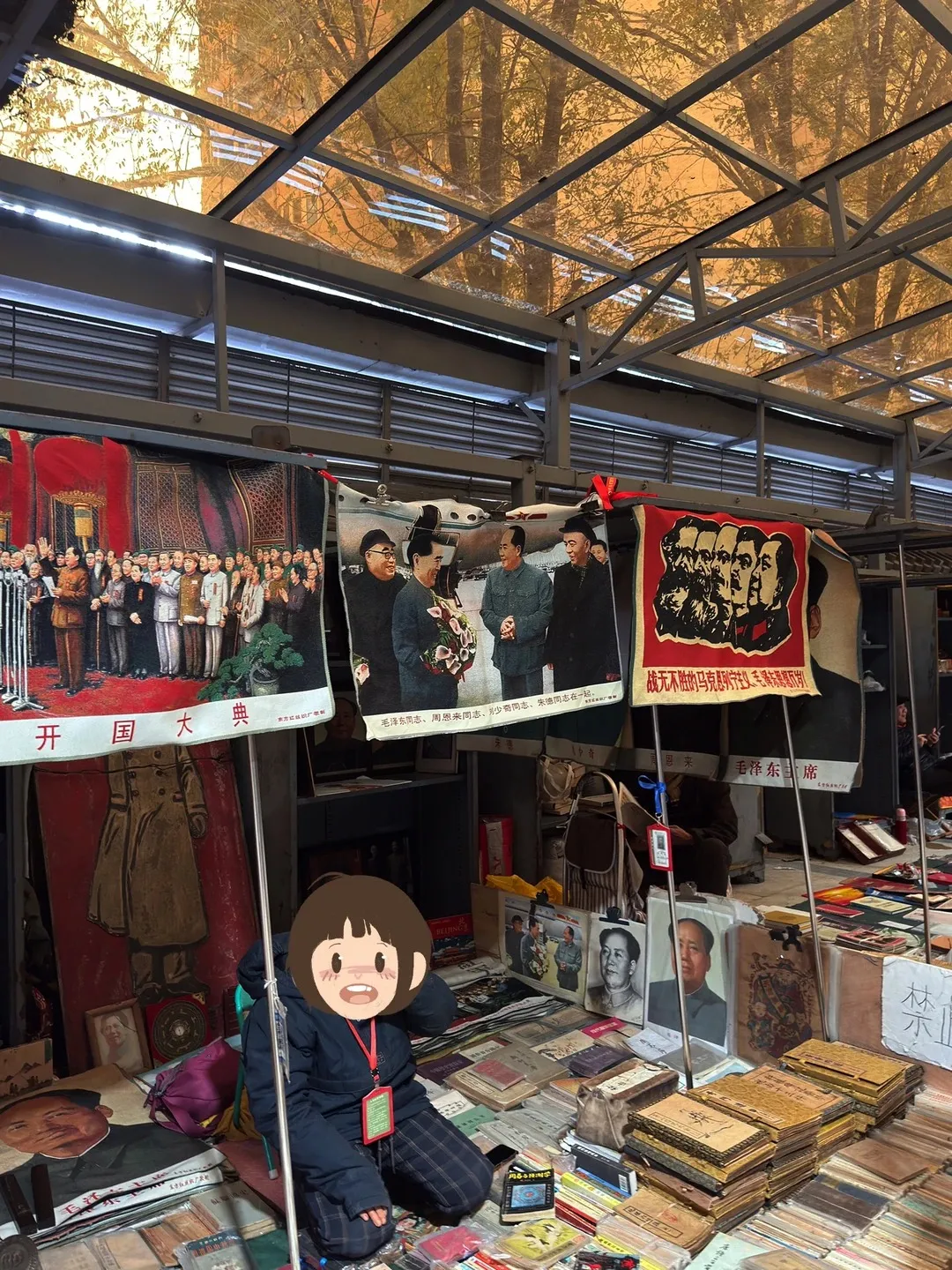 An indoor antique market stall with a vendor kneeling on the ground surrounded by stacks of old books and framed portraits of Mao Zedong. Large vintage tapestries depicting Mao Zedong and other Chinese historical figures hang from the ceiling structure.