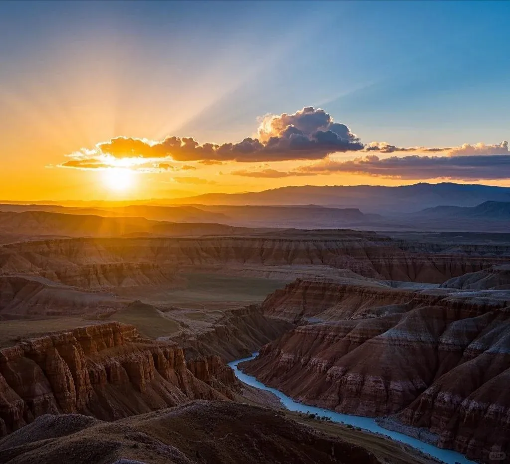 A panoramic view of a deep canyon at sunset, with a bright orange sun on the left horizon casting golden light and rays across the sky. A winding turquoise river flows through the bottom of the canyon, flanked by steep, layered rock walls in shades of brown and red. Distant mountains are silhouetted against the hazy sky.