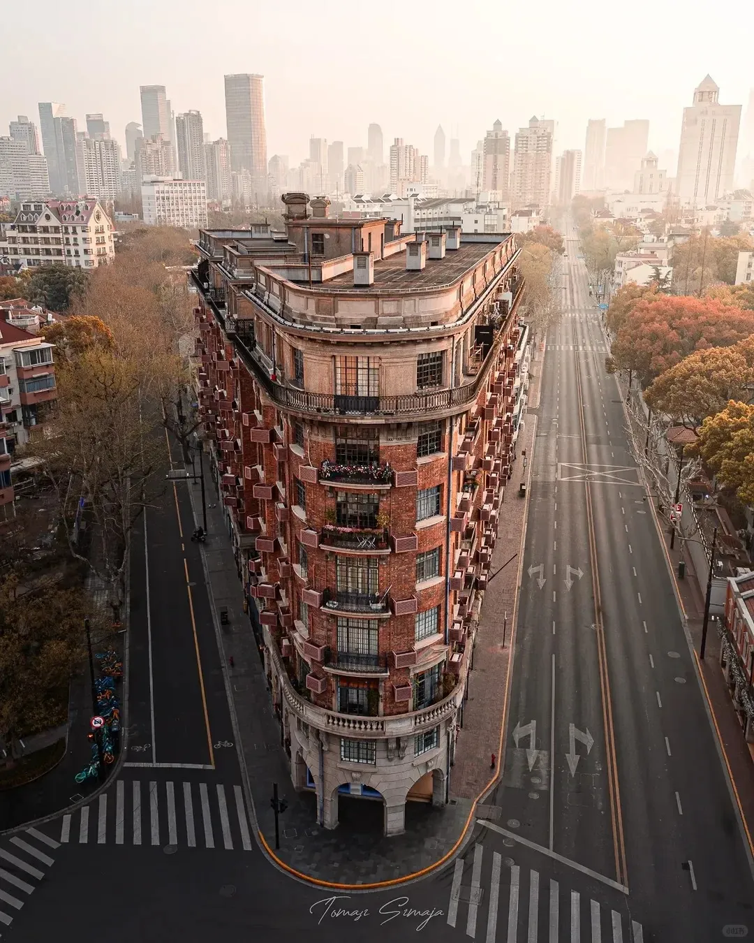 Aerial view of the iconic triangular Wukang Mansion, a red-brick building with rounded balconies, situated at a street intersection in Shanghai's French Concession. Bare autumn trees line the quiet streets, and a hazy city skyline is visible in the distance.