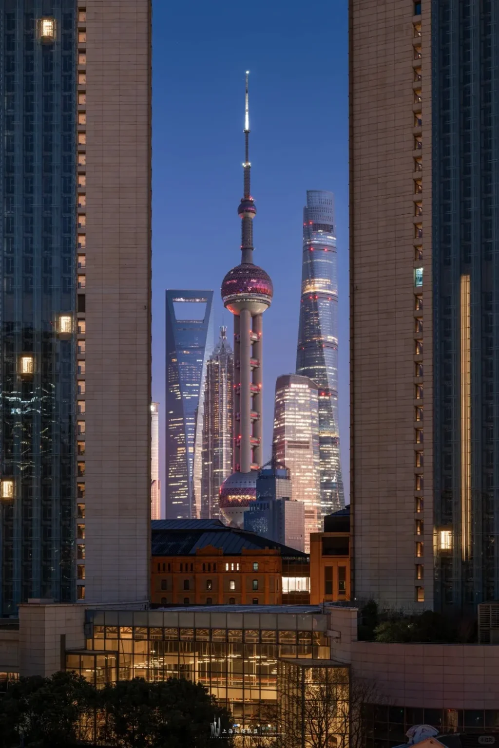 A nighttime view of Shanghai's Pudong skyline, prominently featuring the illuminated Oriental Pearl TV Tower in the center, flanked by the Shanghai World Financial Center on the left and the Shanghai Tower on the right. The iconic skyscrapers are framed by two tall, modern buildings in the immediate foreground, all under a deep blue evening sky.