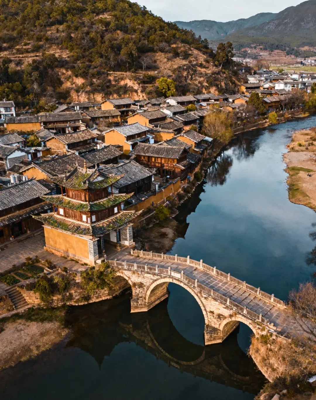 Aerial view of a historic Chinese town featuring a multi-arched stone bridge over a calm river, reflecting the sky and surrounding traditional architecture. Along the riverbank, numerous dark-roofed buildings with light-colored walls line the shore, with a prominent three-tiered temple-like structure standing next to the bridge. Lush, tree-covered mountains rise behind the town under a bright sky.