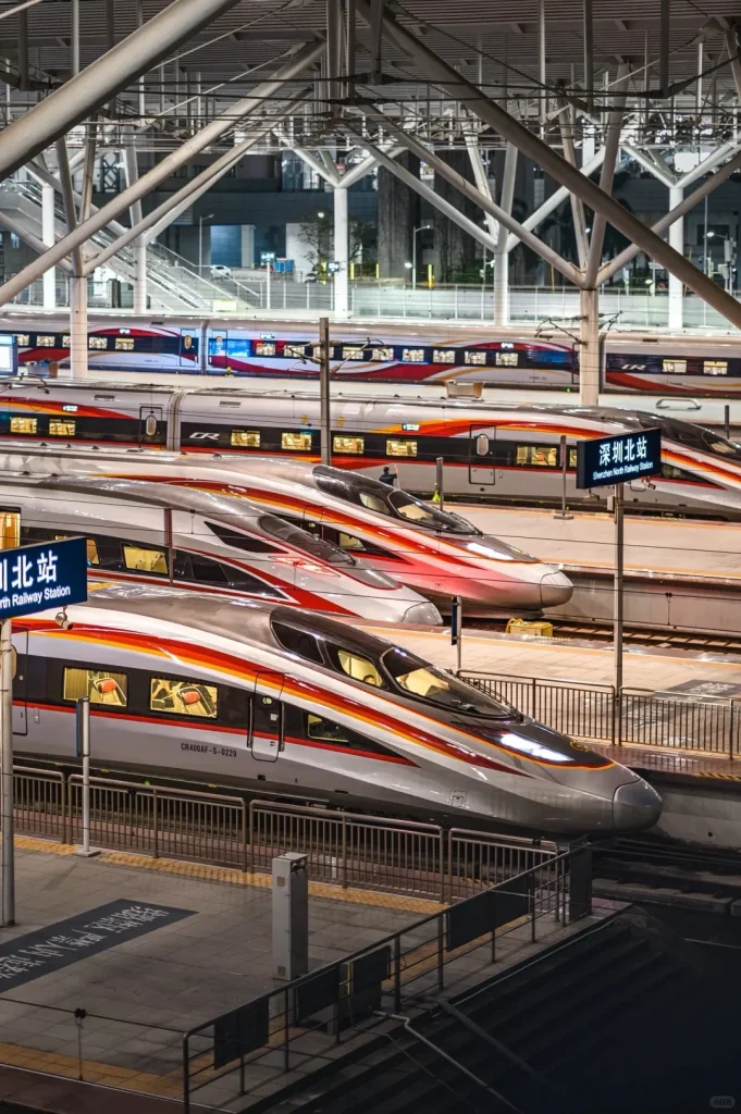 Multiple Fuxinghao high-speed bullet trains, silver with red and orange stripes, are parked at platforms inside Shenzhen North Railway Station at night. Station signs show '深圳北站' and 'Shenzhen North Railway Station'. Overhead metal structures and lighting are visible.
