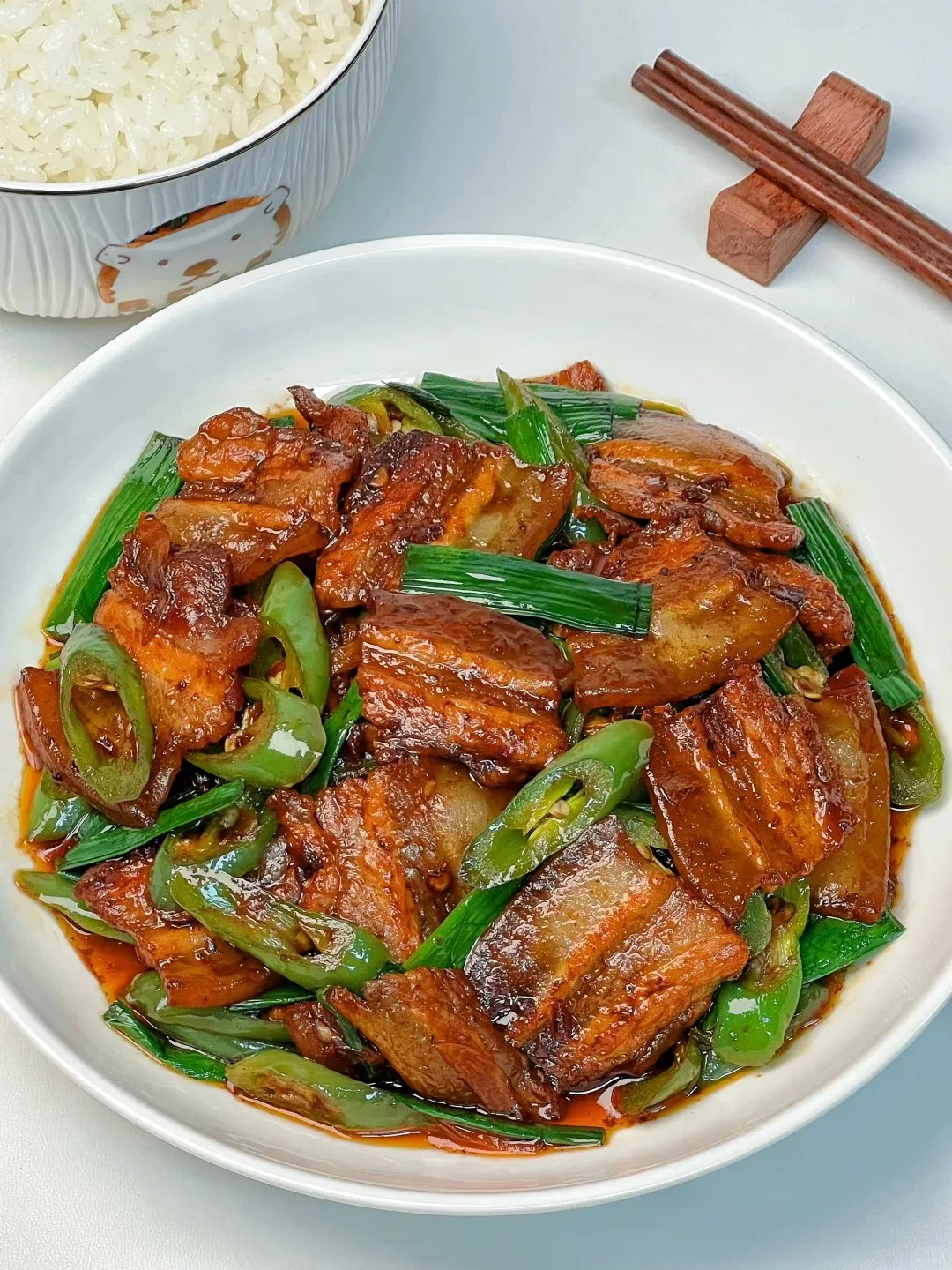 A white plate filled with glistening slices of twice-cooked pork belly, stir-fried with bright green long chili peppers and fresh scallions in a rich, reddish-brown sauce. A bowl of white rice and wooden chopsticks are visible in the background.