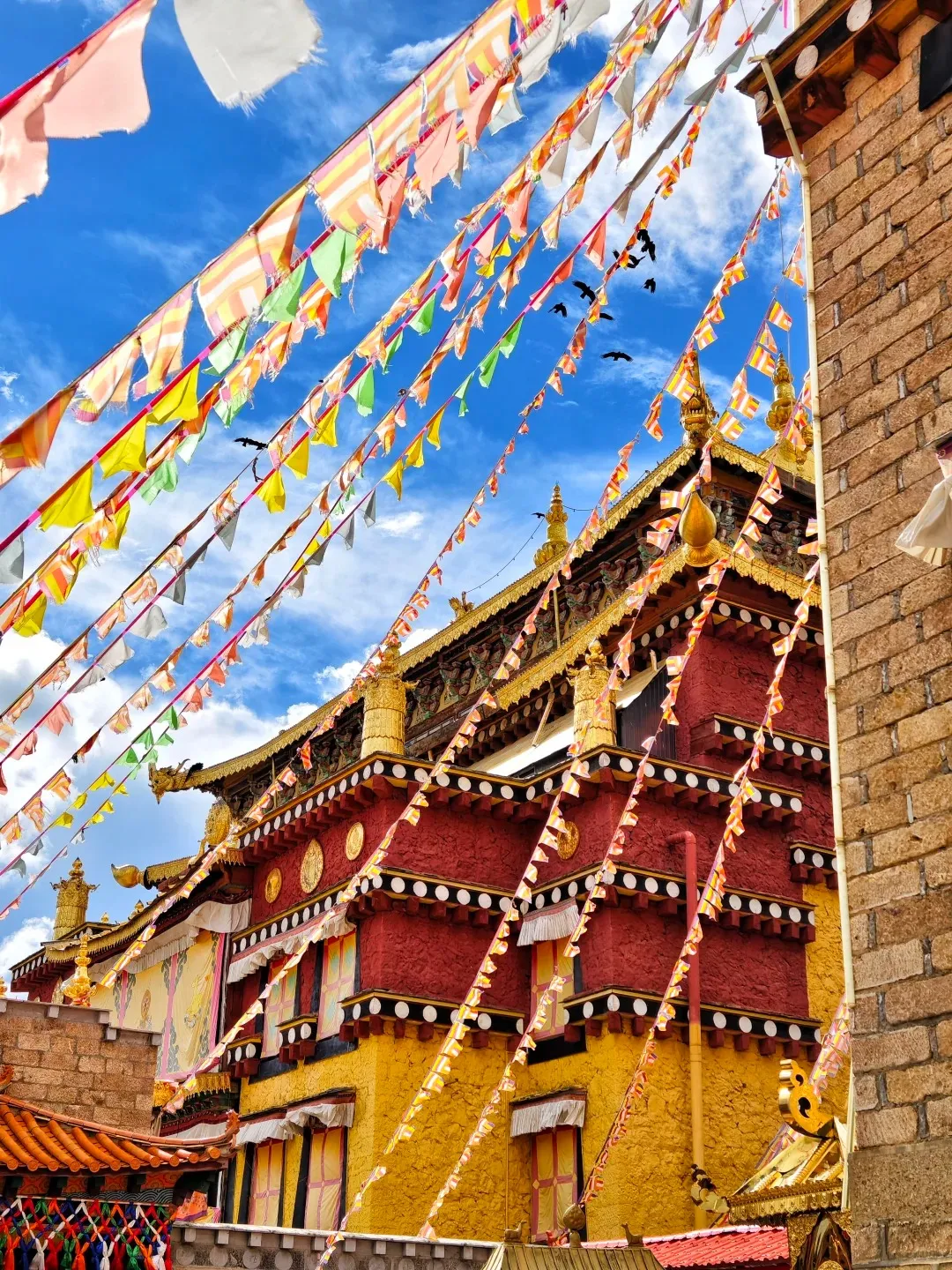 A vibrant red and gold Tibetan Buddhist monastery building with ornate golden roofs under a bright blue sky. Numerous colorful prayer flags are strung diagonally across the image, with several black birds flying amongst them. Parts of a rustic brick wall are visible on the right.