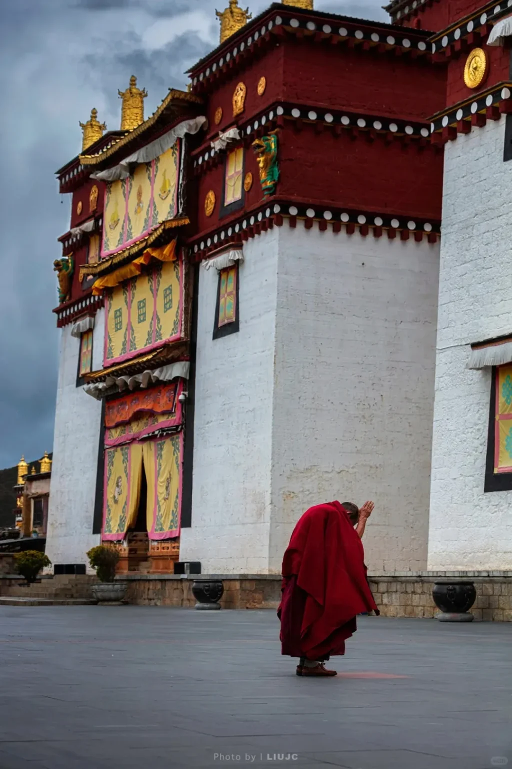 A monk in a voluminous deep red robe is shown from behind, bowing deeply on a large grey paved courtyard. In the background, a multi-story Tibetan monastery building features white lower walls, deep red upper sections with white trim, ornate golden roof decorations, and numerous yellow and pink decorative fabric banners hanging from its windows and doors. The sky is overcast.