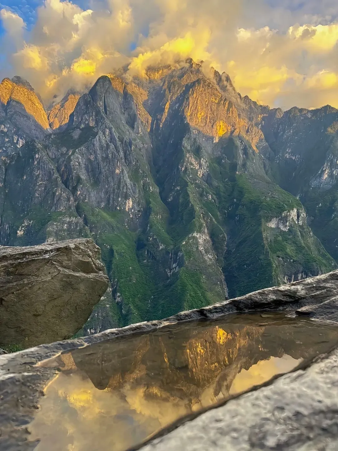 Rugged, steep mountains with green slopes bathed in golden sunset light and clouds, reflected in a puddle on a rock in the foreground.