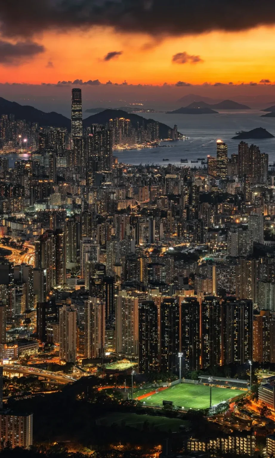 Panoramic view of Hong Kong city skyline at dusk from Victoria Peak. The sky is an orange and dark grey gradient above illuminated skyscrapers. Victoria Harbour with islands is in the distance, and a brightly lit football field is in the foreground among dense buildings.