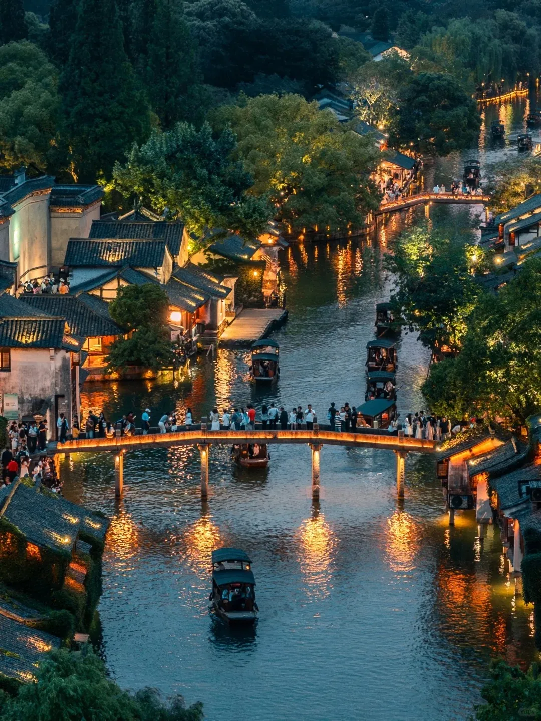 High-angle night view of Wuzhen West Scenic Zone, showing a winding canal illuminated by warm lights reflecting on the water. Traditional Chinese buildings with dark tiled roofs line the canal banks. A stone arch bridge with many people crosses the canal, and several traditional wooden boats with passengers are visible on the water. Lush green trees surround the area.